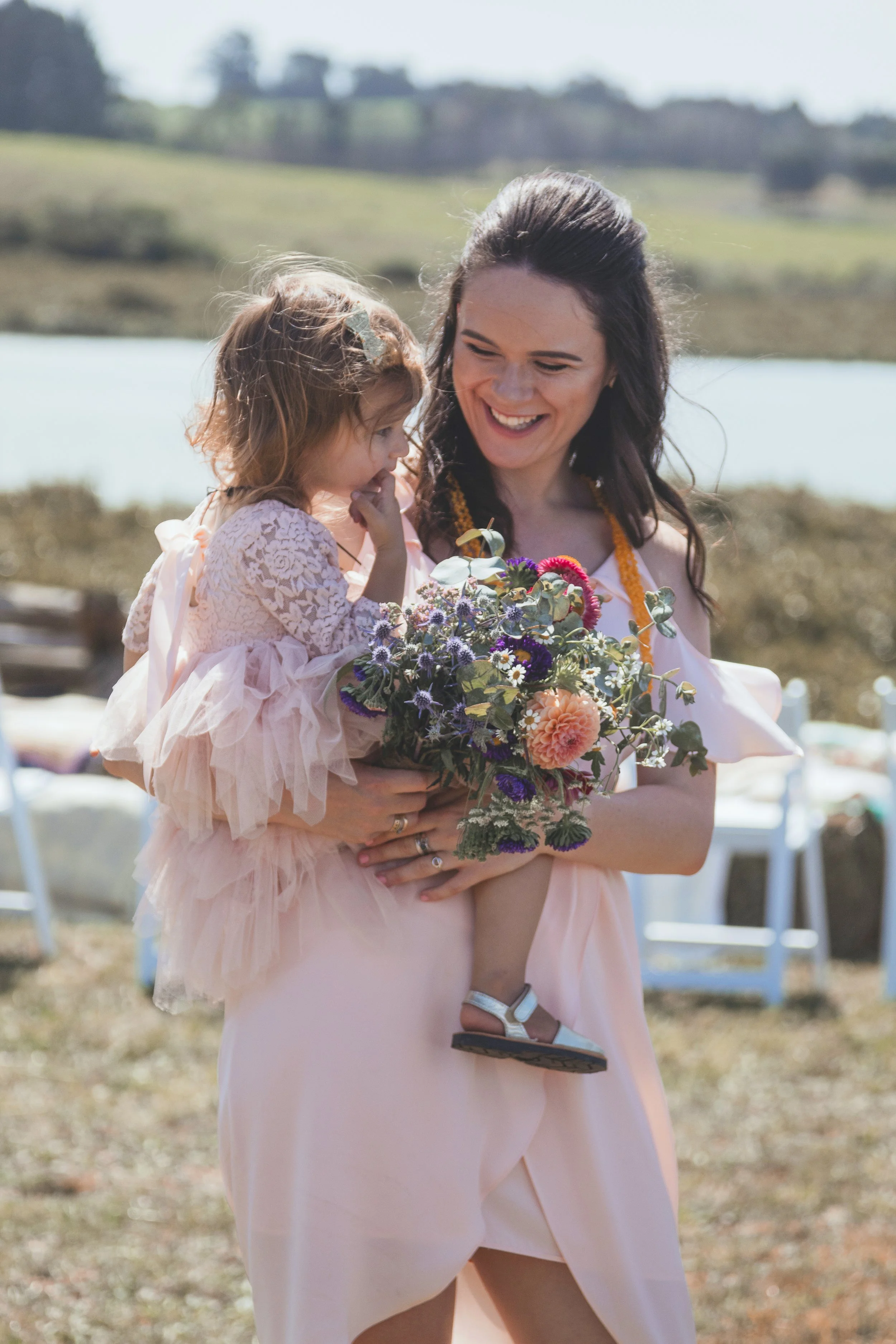 A woman in a pink dress holding a young girl in a pink dress and flowers, outdoors with a lake and hills in the background.
