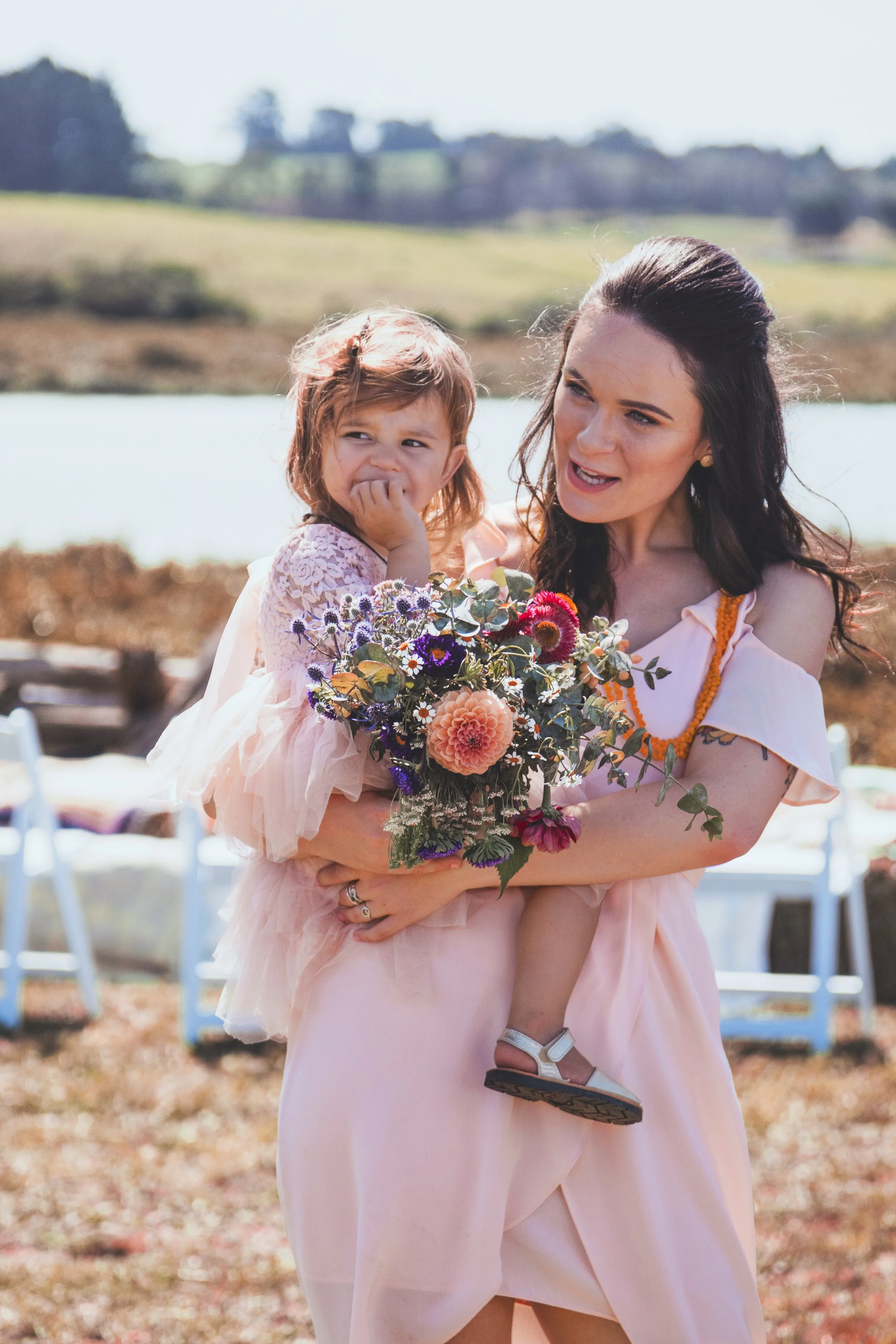 A woman with dark hair and a young girl with light brown hair in a pink dress are outdoors, with the woman holding the girl and a bouquet of colorful flowers, in a setting with chairs and a scenic landscape in the background.