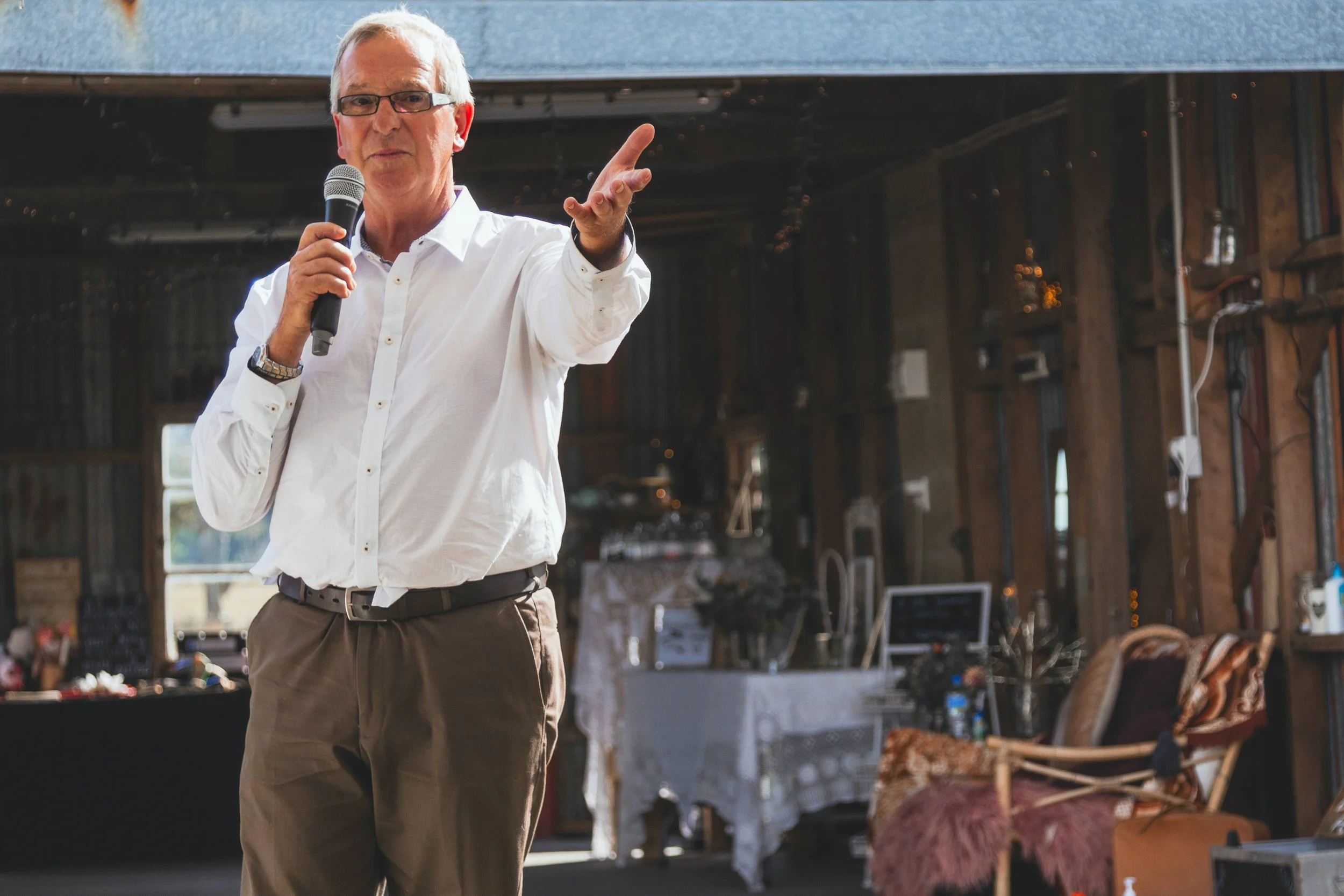 An older man with glasses, wearing a white shirt, speaking into a microphone and gesturing with his hand, inside a rustic building with string lights and wooden furnishings.