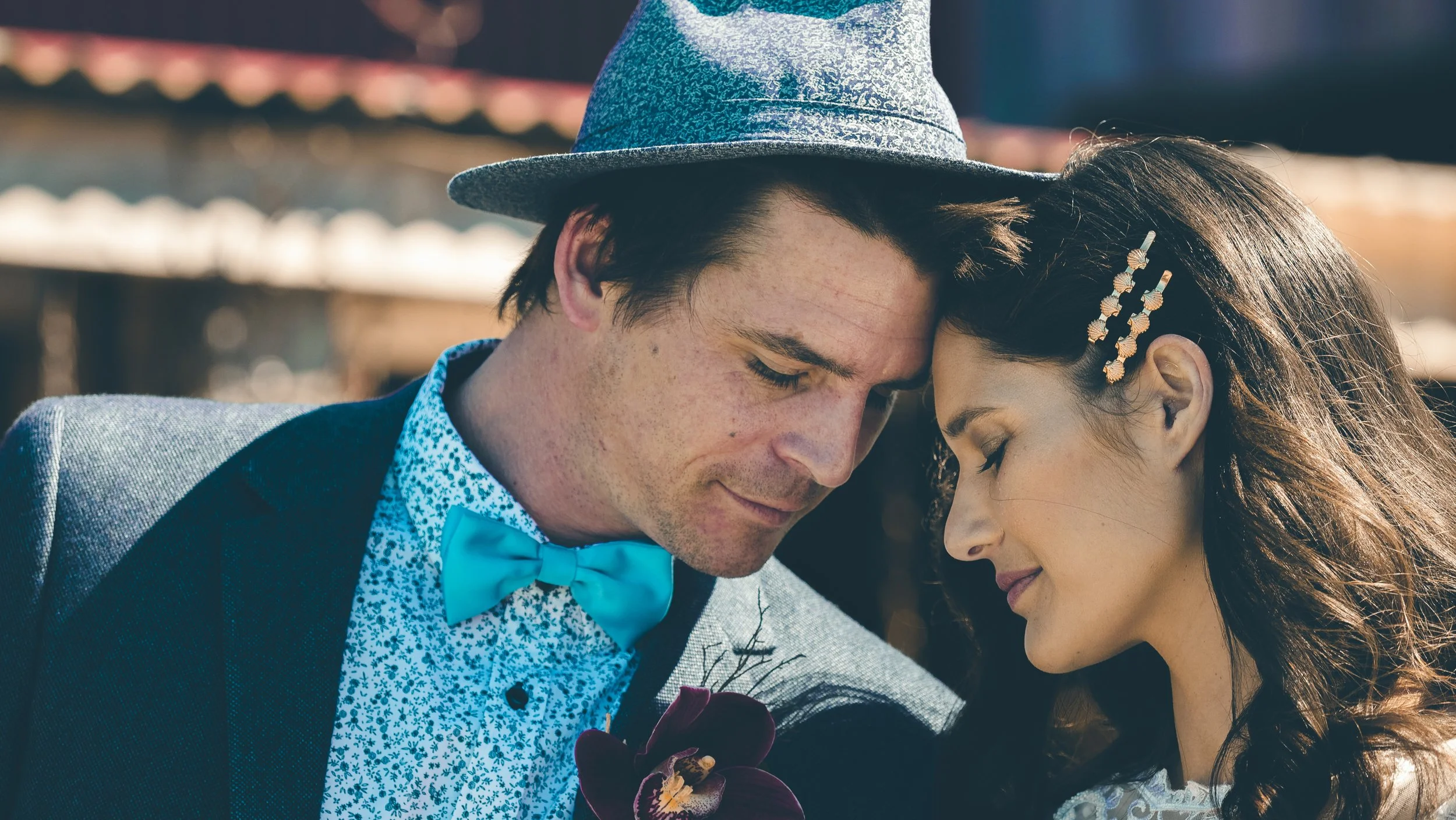 A close-up of a man and woman with their foreheads touching, eyes closed, sharing an intimate moment. The man is wearing a gray fedora hat, a blue bow tie, and a patterned shirt, while the woman has long brown hair with decorative hair clips and is w