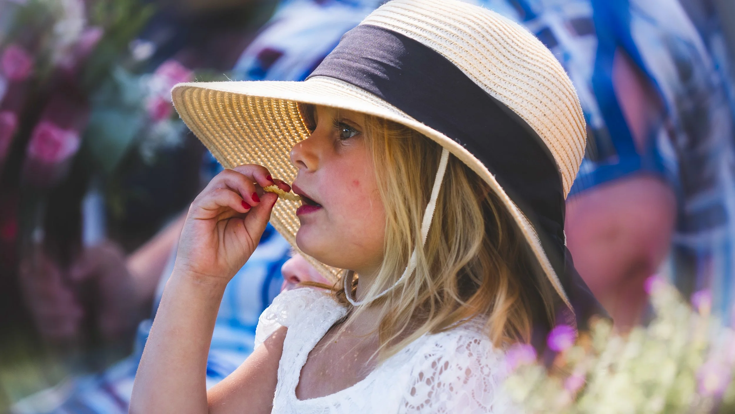 A young girl wearing a large straw sun hat with a black ribbon, holding a cookie near her mouth, in an outdoor garden setting.