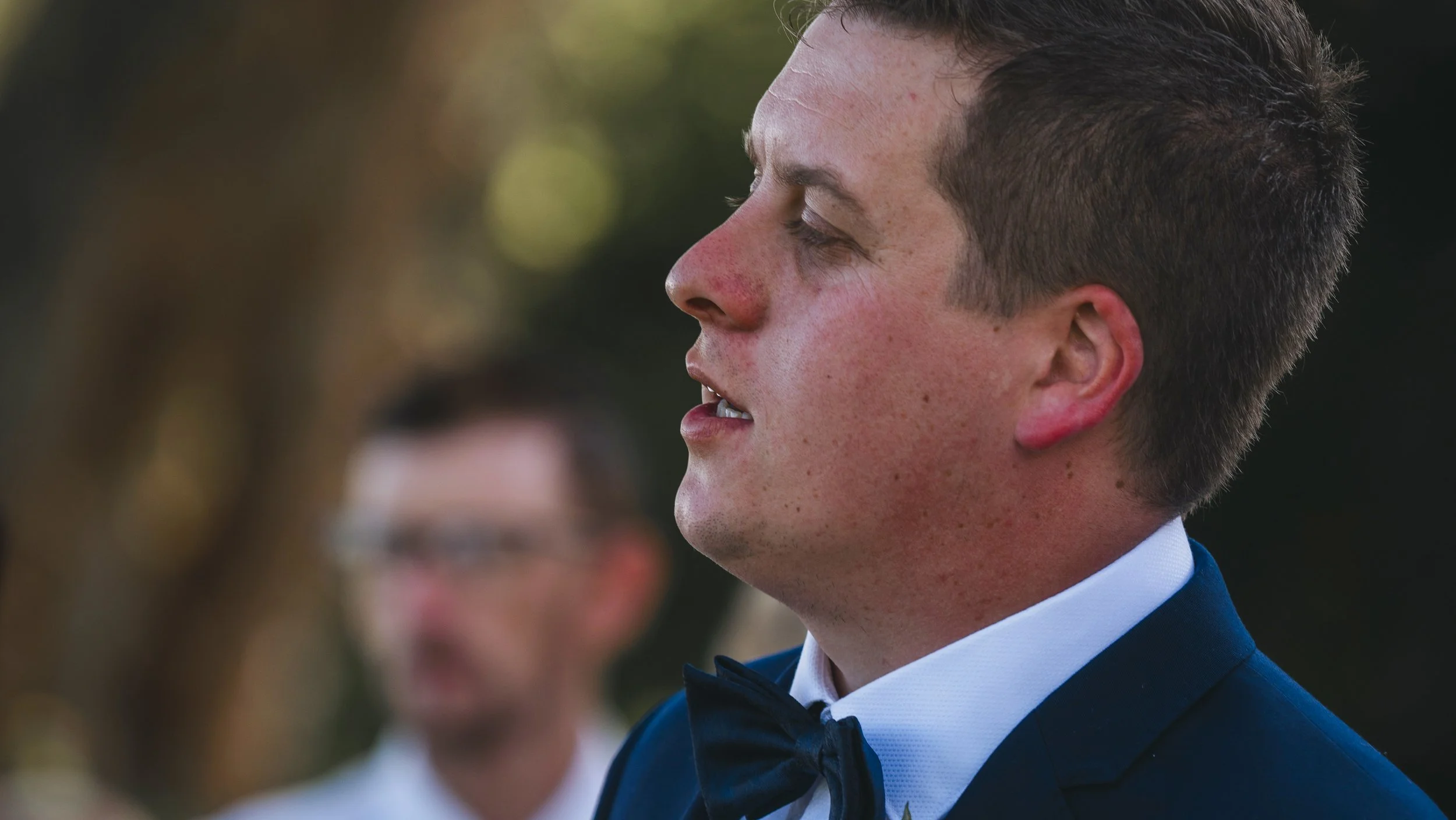 A young man with short dark hair, wearing a blue tuxedo with a black bow tie, standing outdoors during sunset, with a blurred woman in the background.