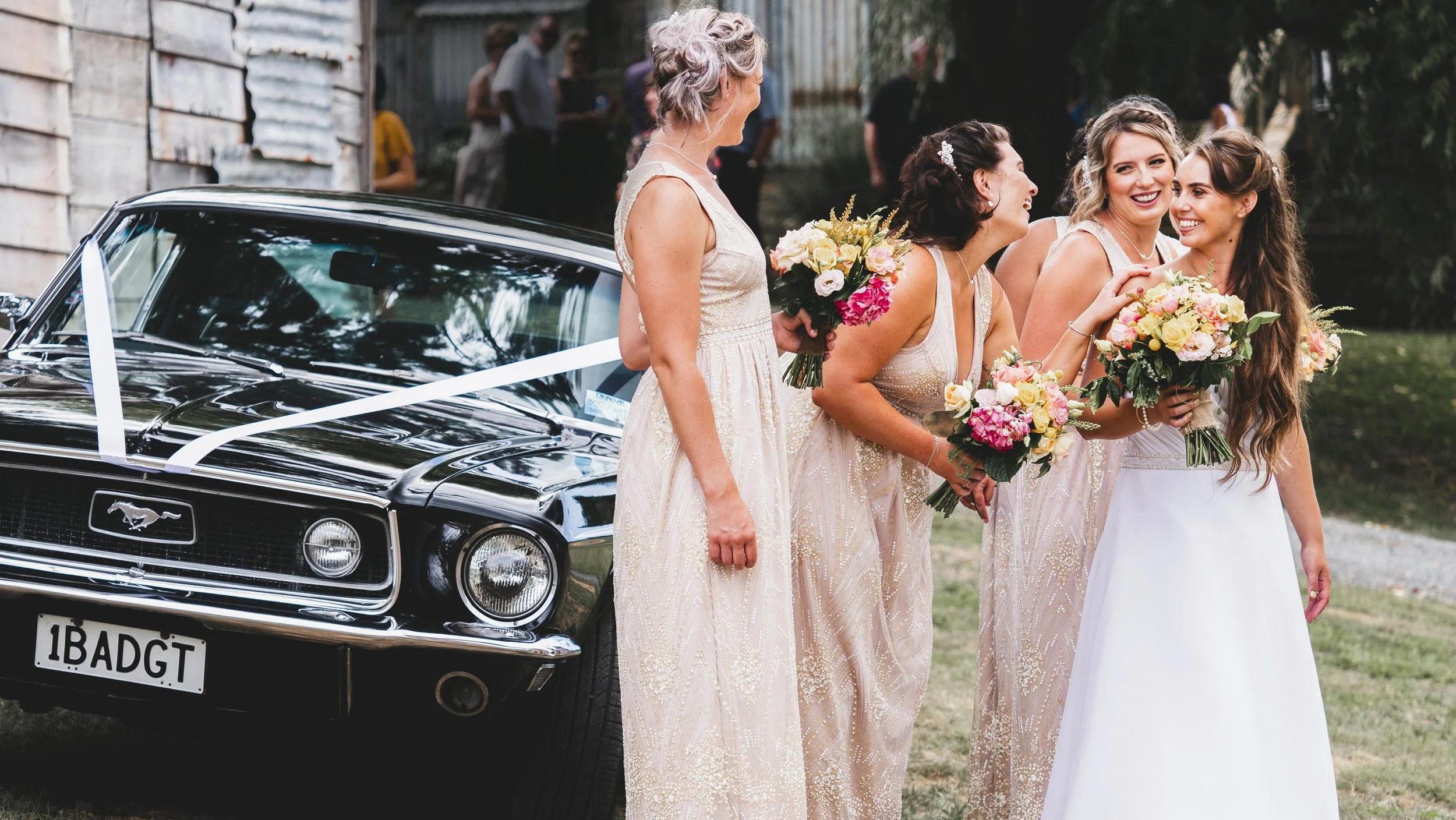 Women in bridesmaid dresses holding bouquets standing near a black vintage Ford Mustang with a white ribbon, at an outdoor wedding celebration.