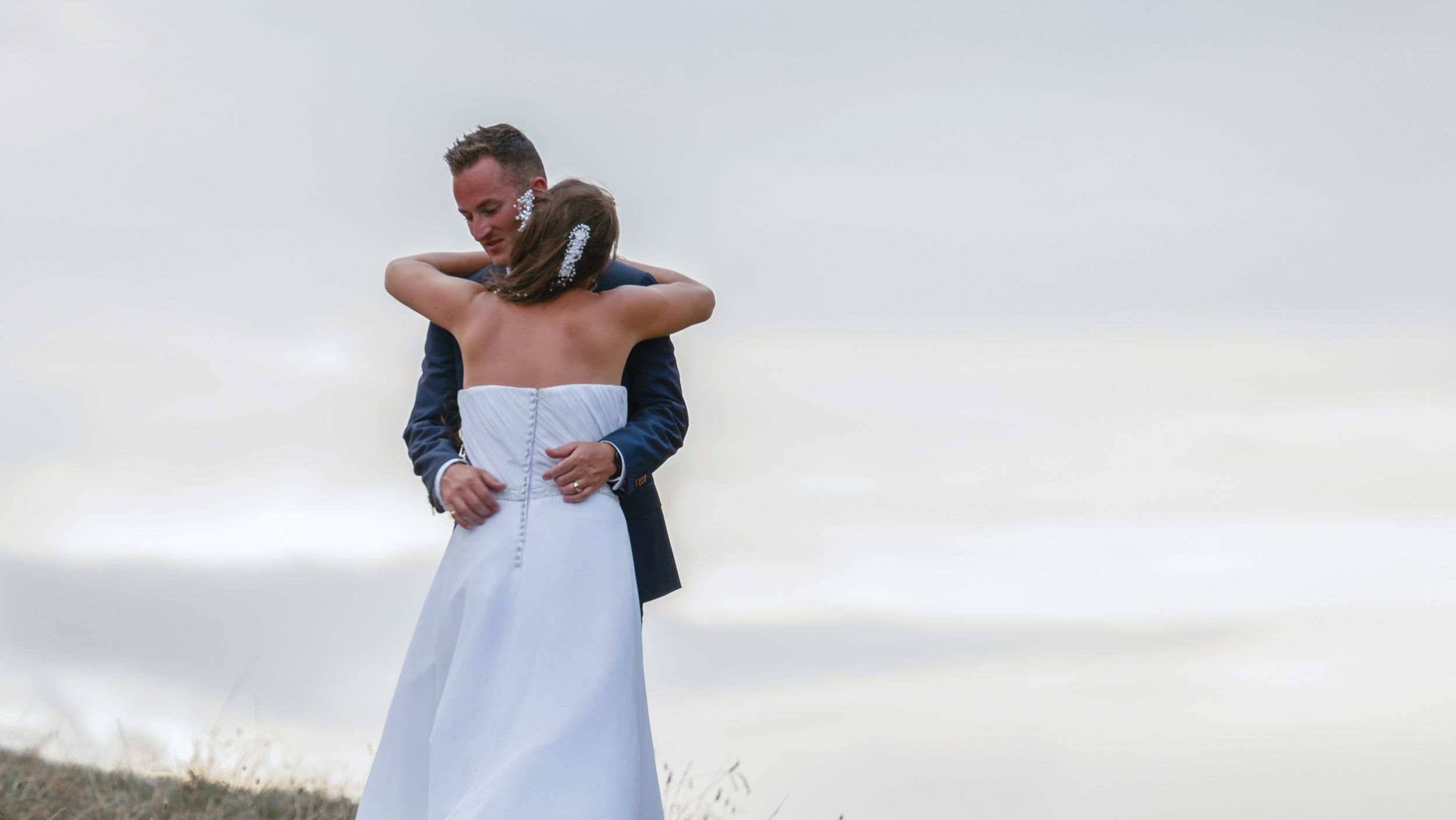 A man and woman embracing outdoors, possibly during a wedding or special event, with a cloudy sky in the background.