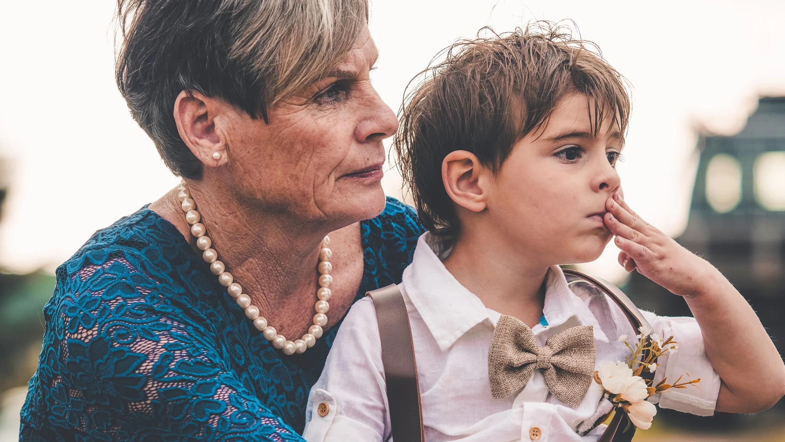 A woman with short brown hair and a pearl necklace, wearing a blue patterned dress, standing beside a young boy with brown hair, a white shirt, and a beige bow tie, holding flowers, with a blurred outdoor background.