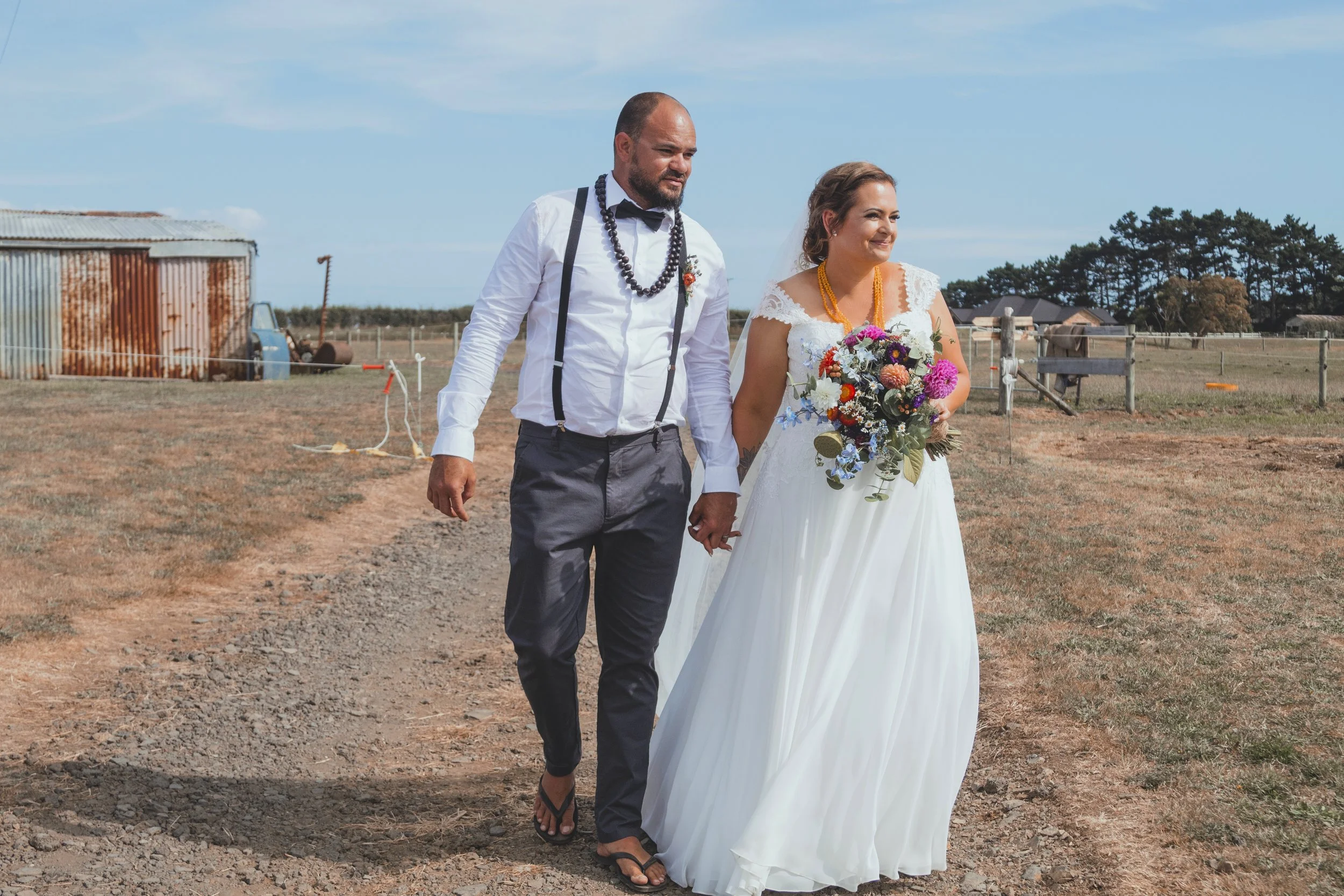 A man and a woman holding hands outdoors during a wedding, with the man wearing a white shirt, suspenders, a bow tie, and beaded necklaces, and the woman wearing a white wedding dress and holding a bouquet of flowers.