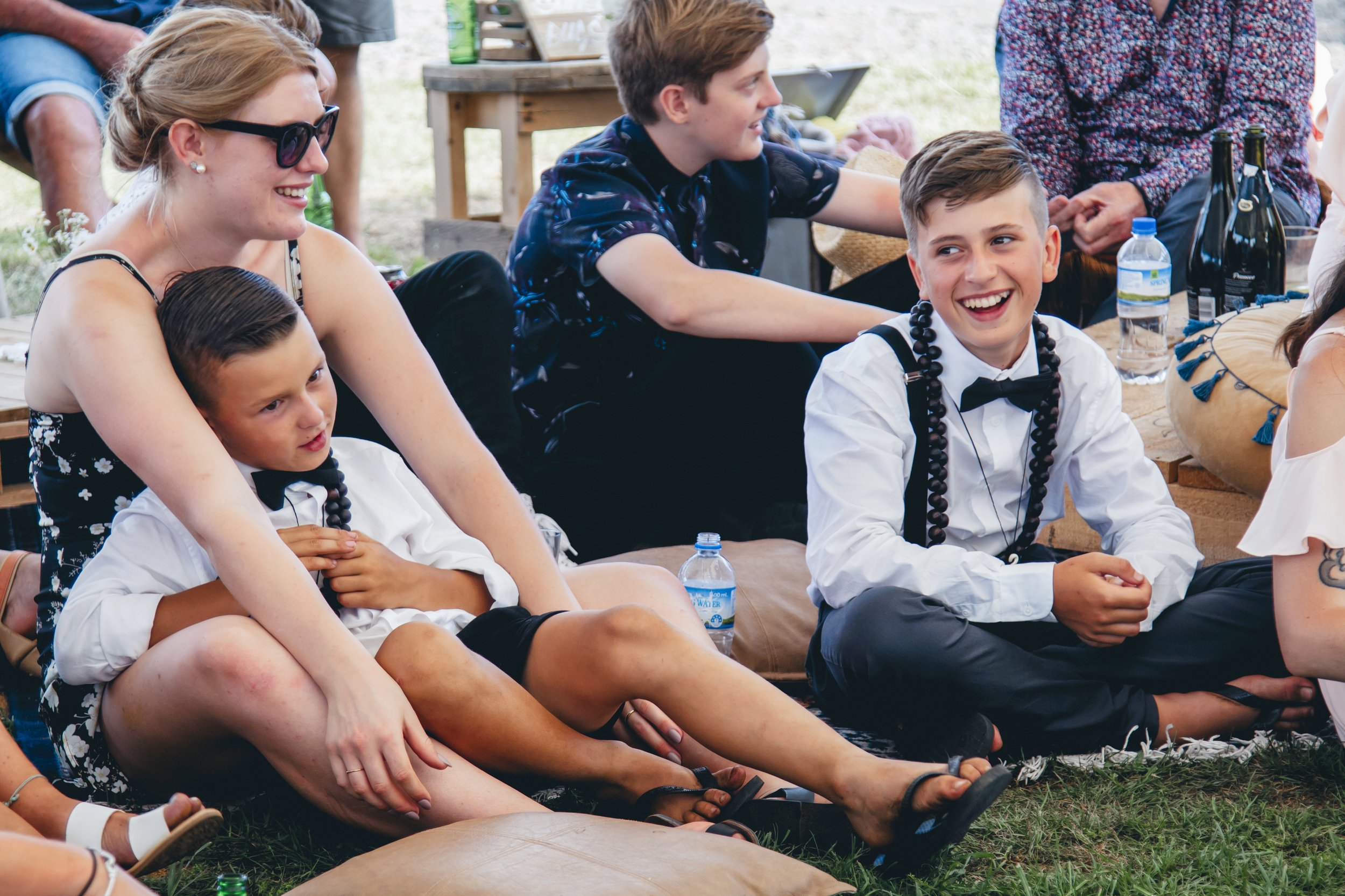 Group of young people sitting on the grass at an outdoor event, smiling and enjoying each other's company. Some are dressed casually, while others are dressed more formally with bow ties.