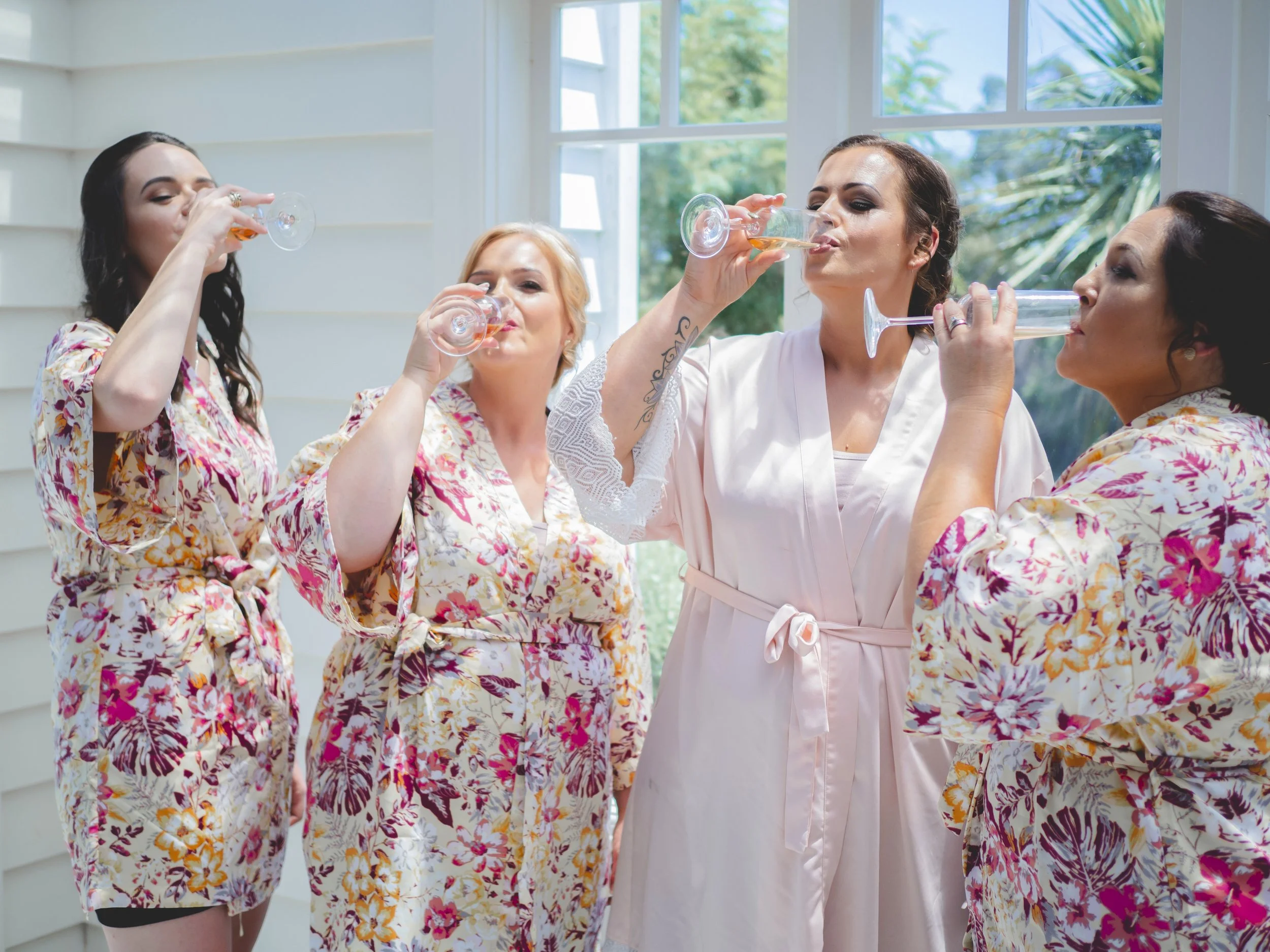 Four women in floral robes drinking wine on a patio with large windows and trees outside.