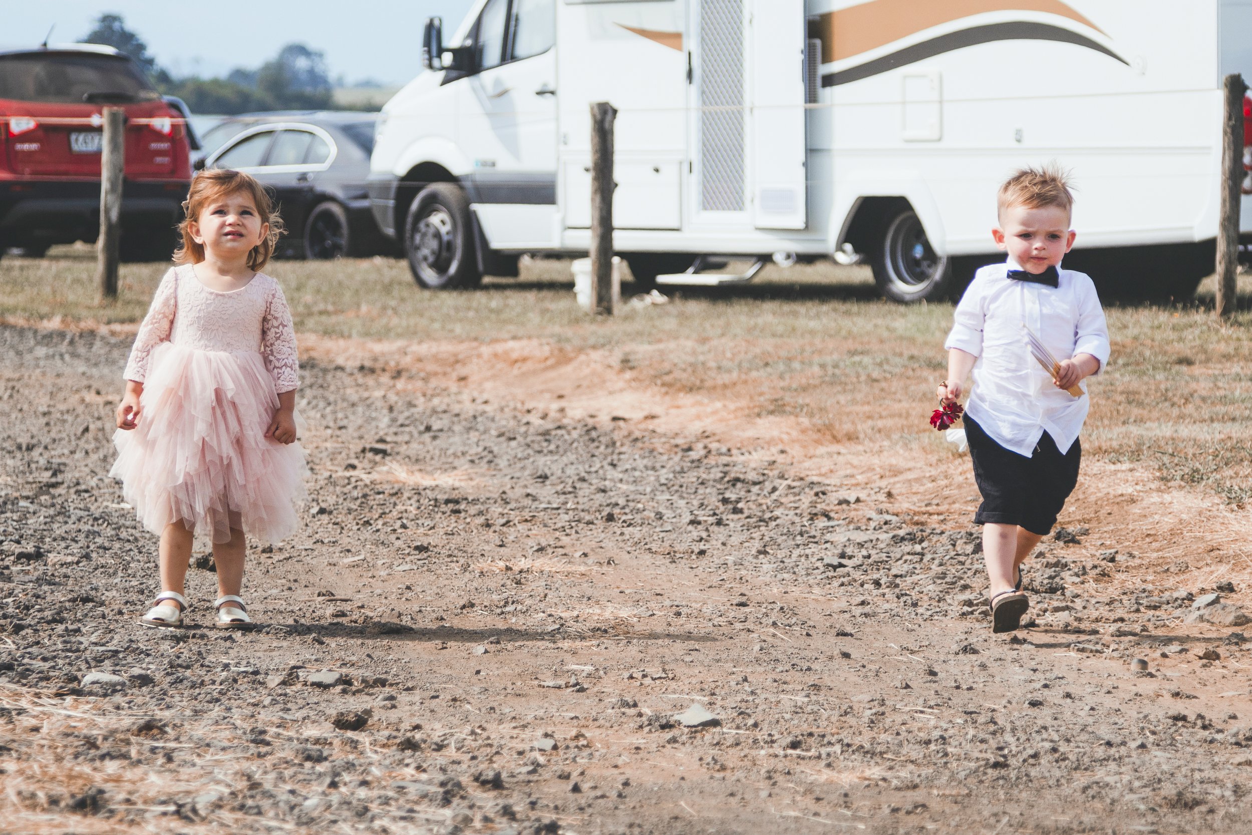 Young girl in a pink dress standing on a dirt path, and a young boy in a white shirt and black shorts walking with a small bouquet of flowers, with cars and a camper in the background.