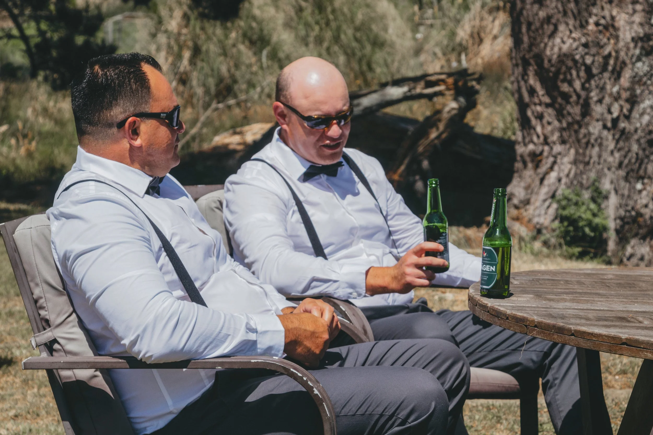 Two men dressed in white shirts with suspenders and black bow ties sitting outdoors at a round wooden table, holding green glass bottles, with trees in the background.