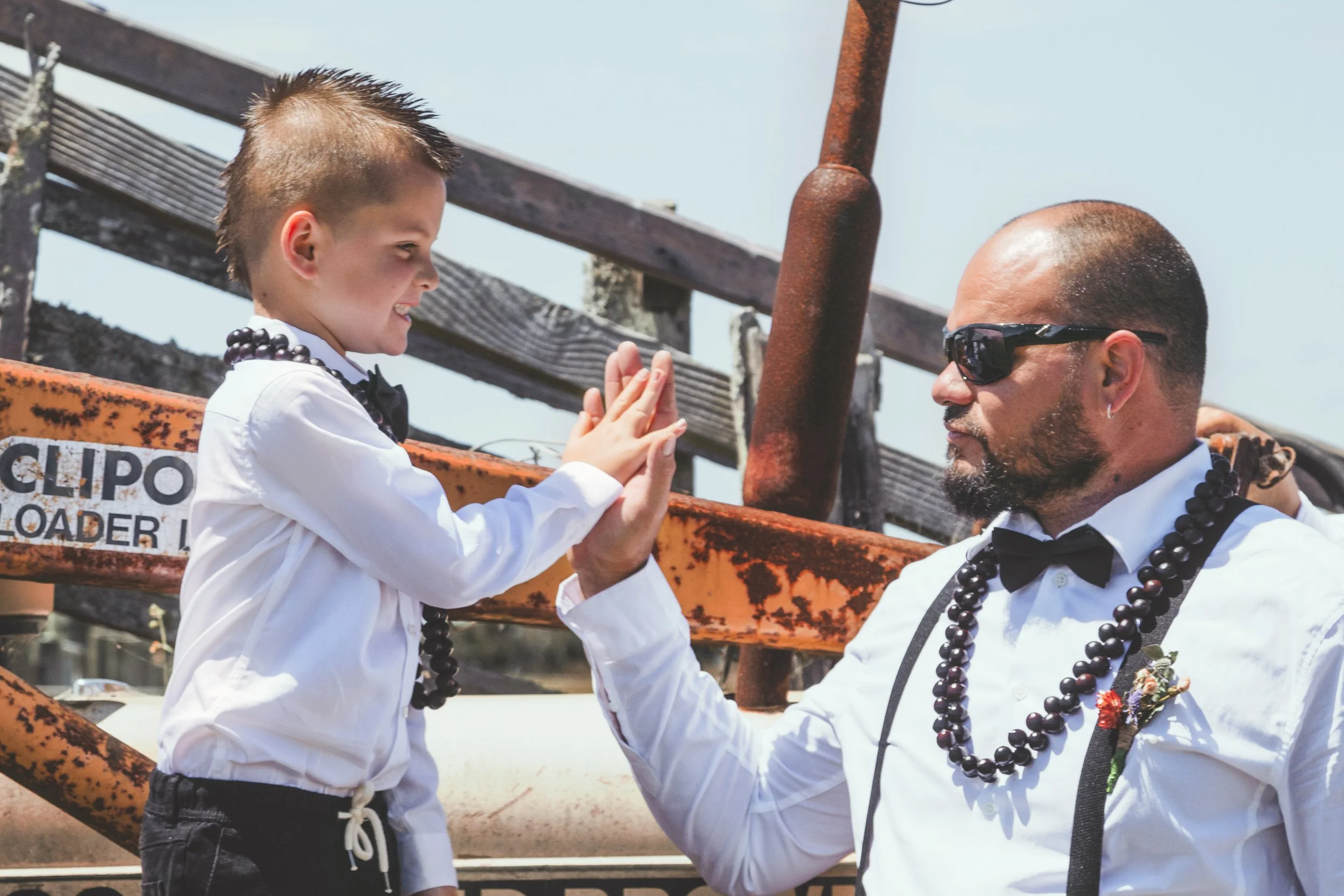 A young boy and a man dressed in formal attire giving each other a high five outdoors. The boy is wearing a white shirt, black bow tie, dark pants, and a beaded necklace. The man is wearing sunglasses, a white shirt, black suspenders, a black bow tie