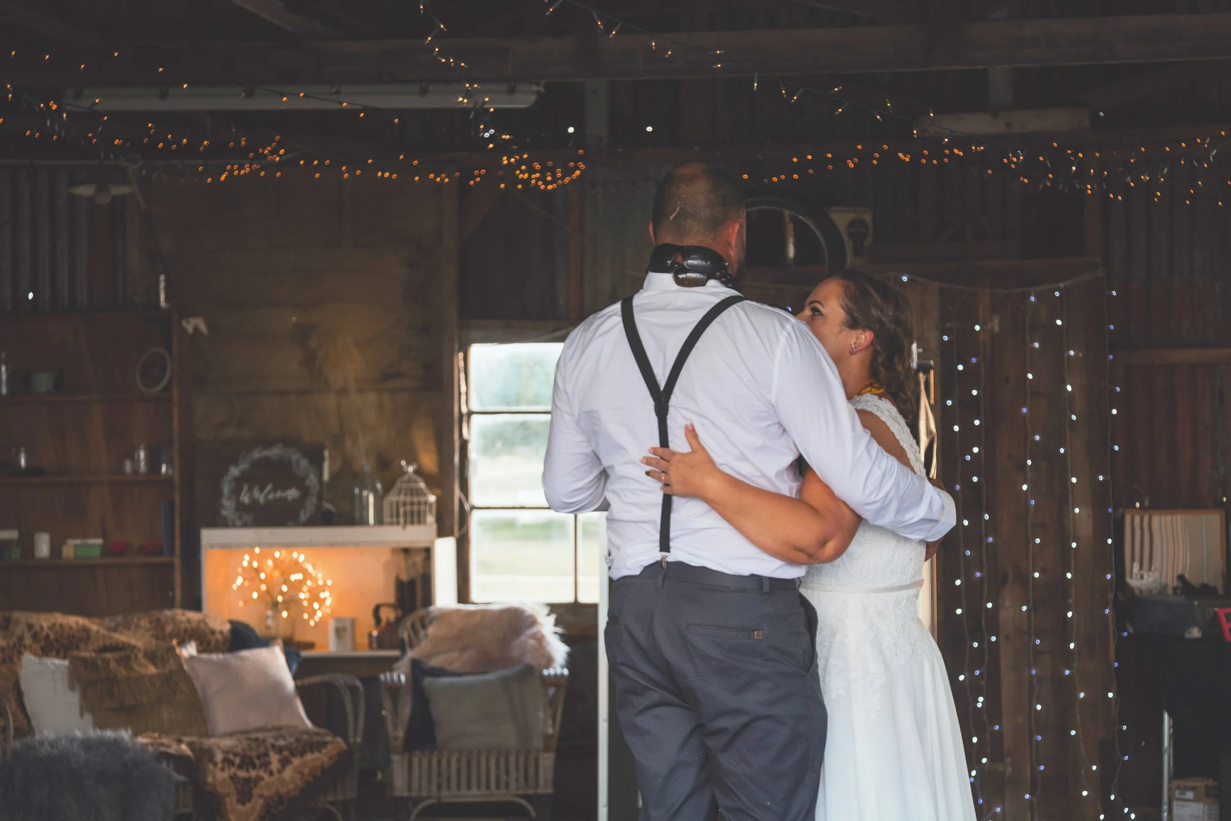 A couple dancing closely inside a rustic barn decorated with string lights for a wedding.