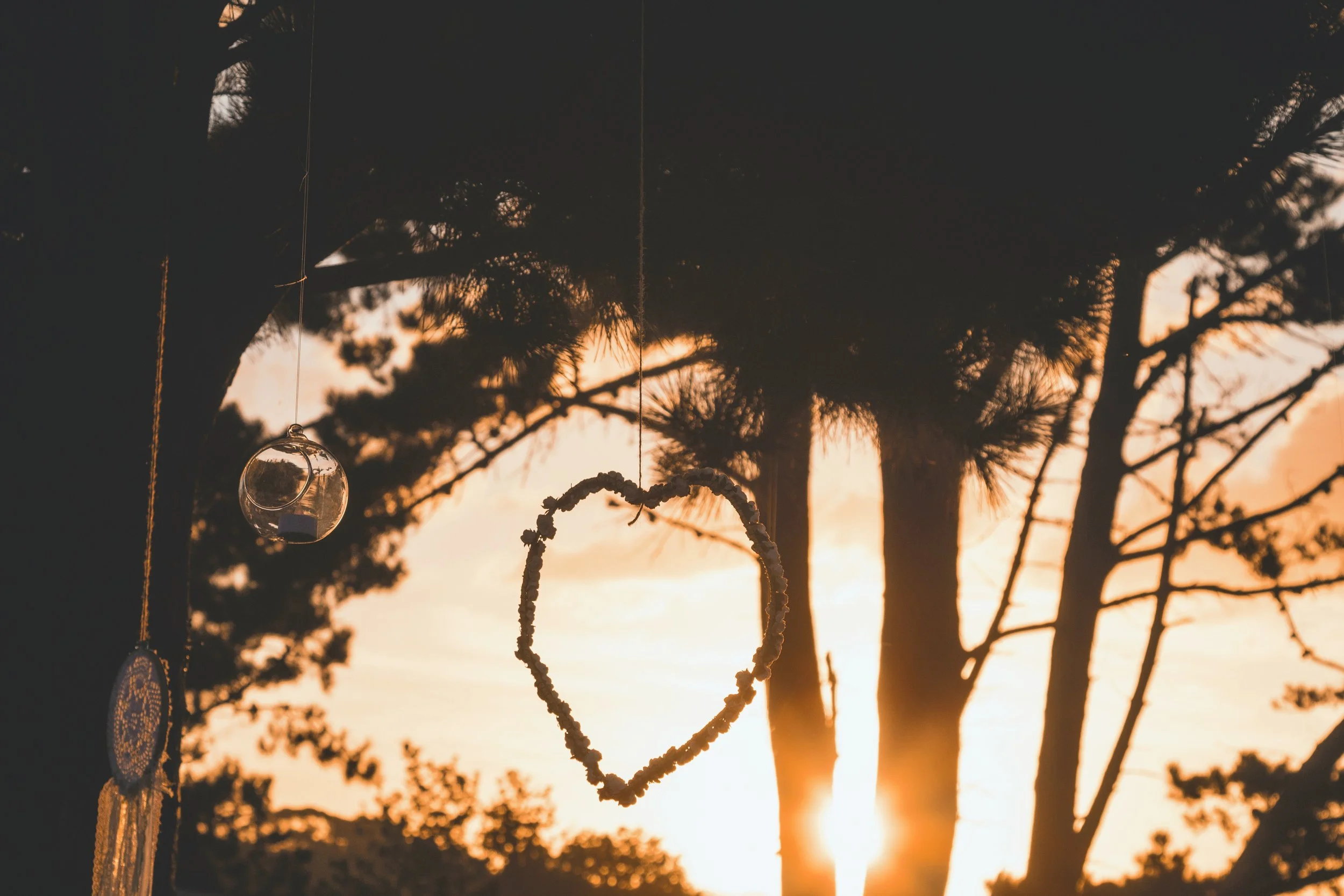Decorative hanging ornaments, including a clear glass orb and a heart-shaped wreath, hanging in front of silhouette trees at sunset.