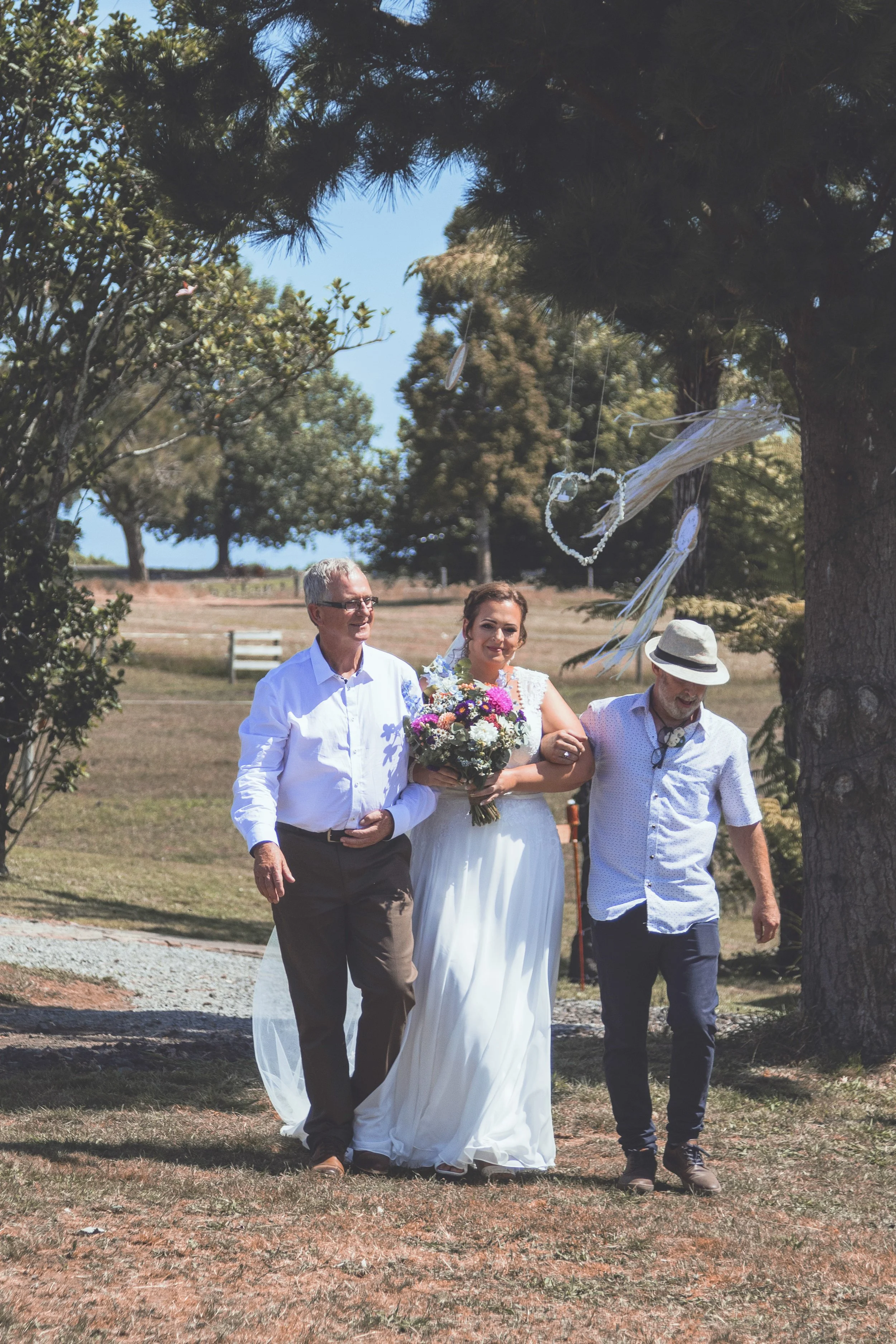 A bride in a white wedding dress holding a bouquet of flowers walks outdoors with two men, one in a white shirt and khakis, and the other in a light blue shirt and dark pants. They are walking under a large tree with wind chimes hanging from its bran