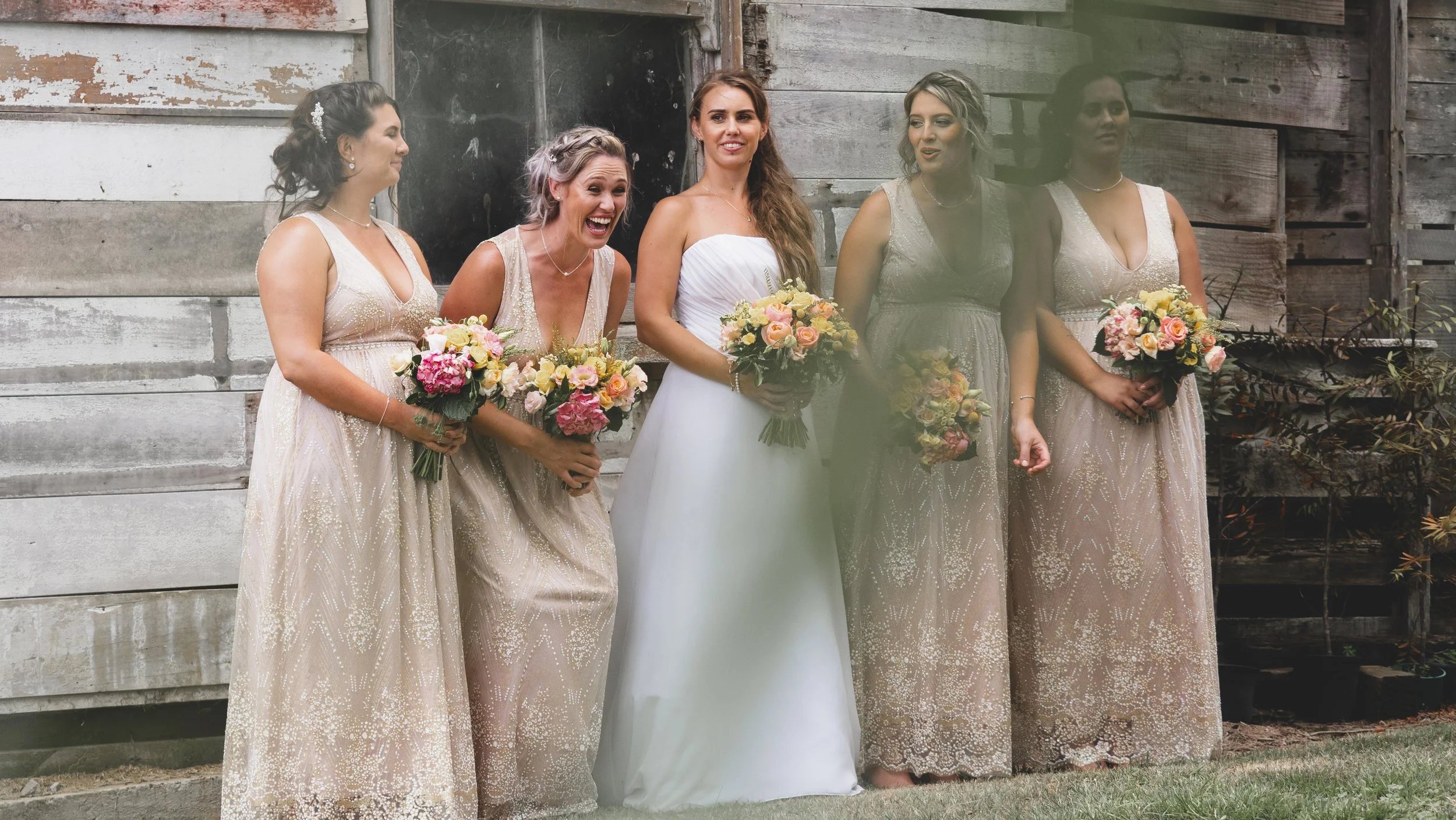 A group of women, including a bride in a white wedding gown holding a bouquet, and four bridesmaids in matching light beige dresses holding bouquets, standing outdoors in front of a rustic wooden wall.
