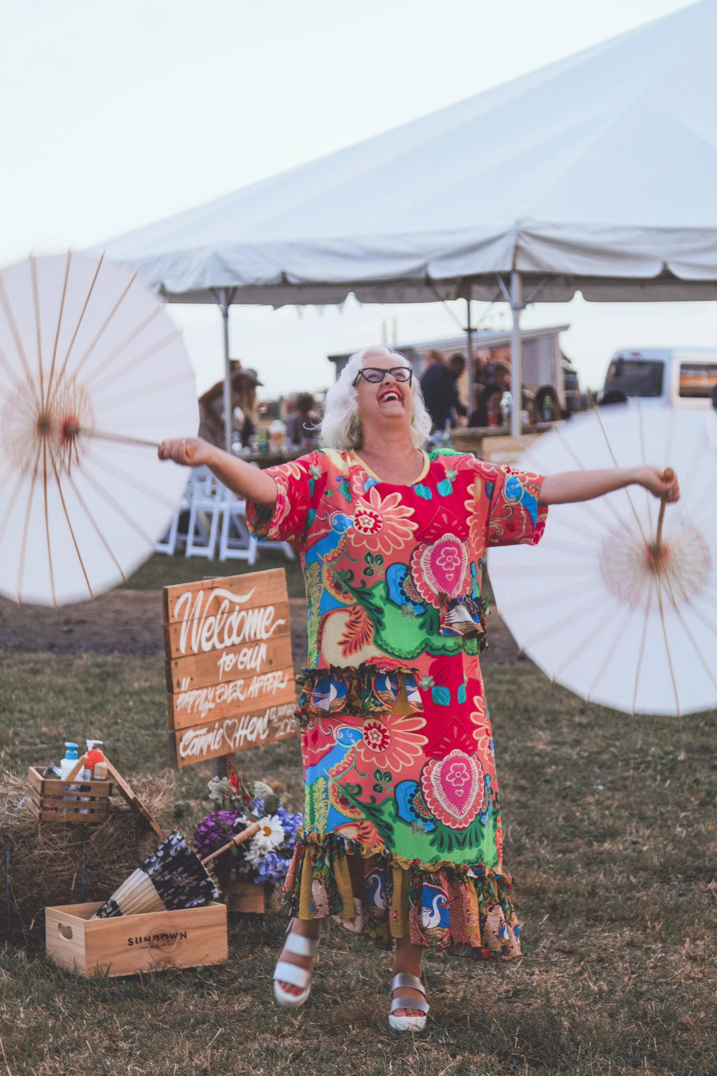 A woman in a colorful floral dress with her arms outstretched, holding paper umbrellas, smiling at an outdoor event under a large white canopy with people and tables in the background.