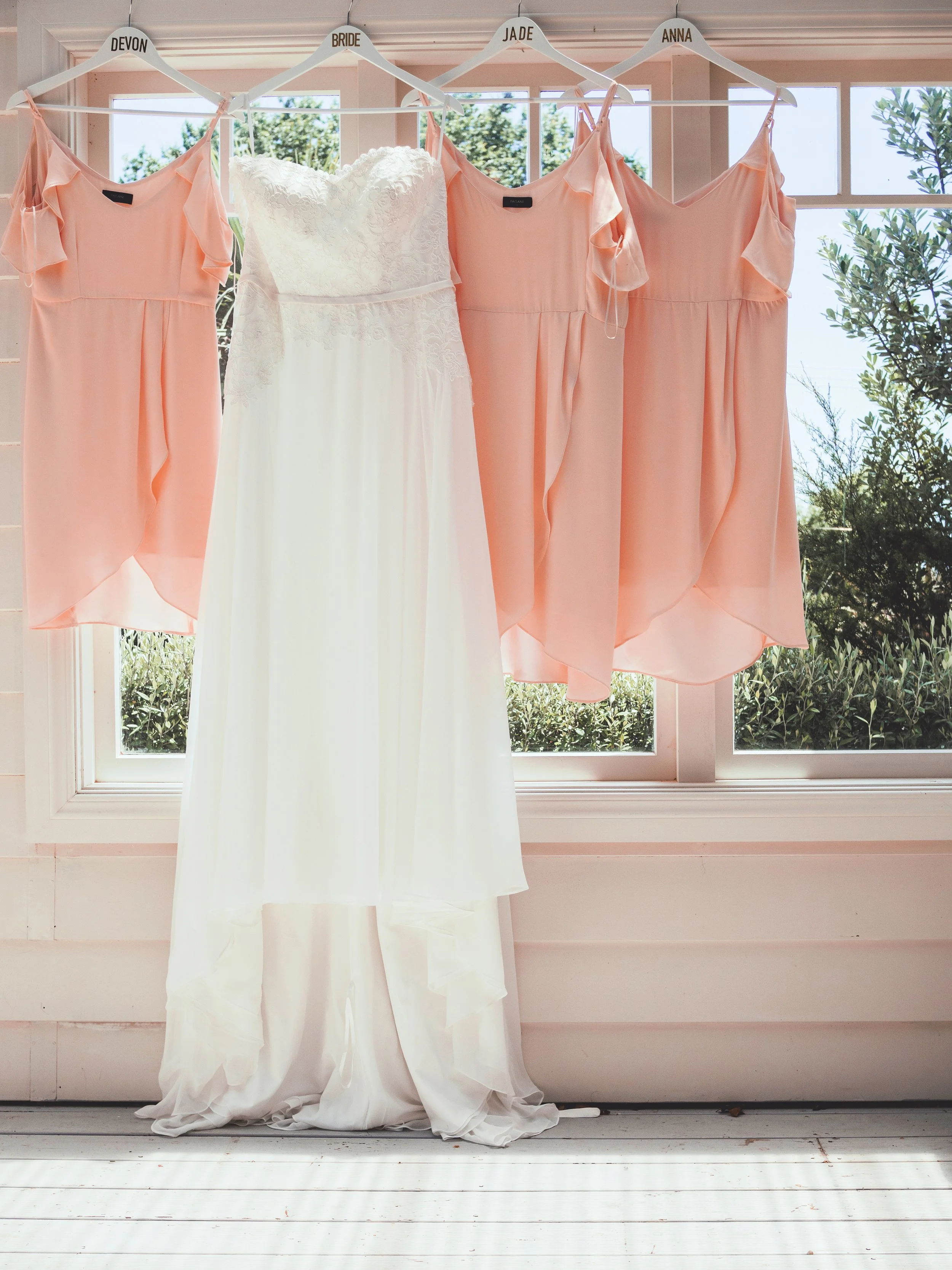 Wedding dresses and bridesmaid dresses hanging on a clothesline outside. The dresses are labeled with hangers that read 'Devon,' 'Bride,' 'Jade,' and 'Anna.' The wedding dress is white with lace details, and the bridesmaid dresses are light peach with ruffled sleeves.