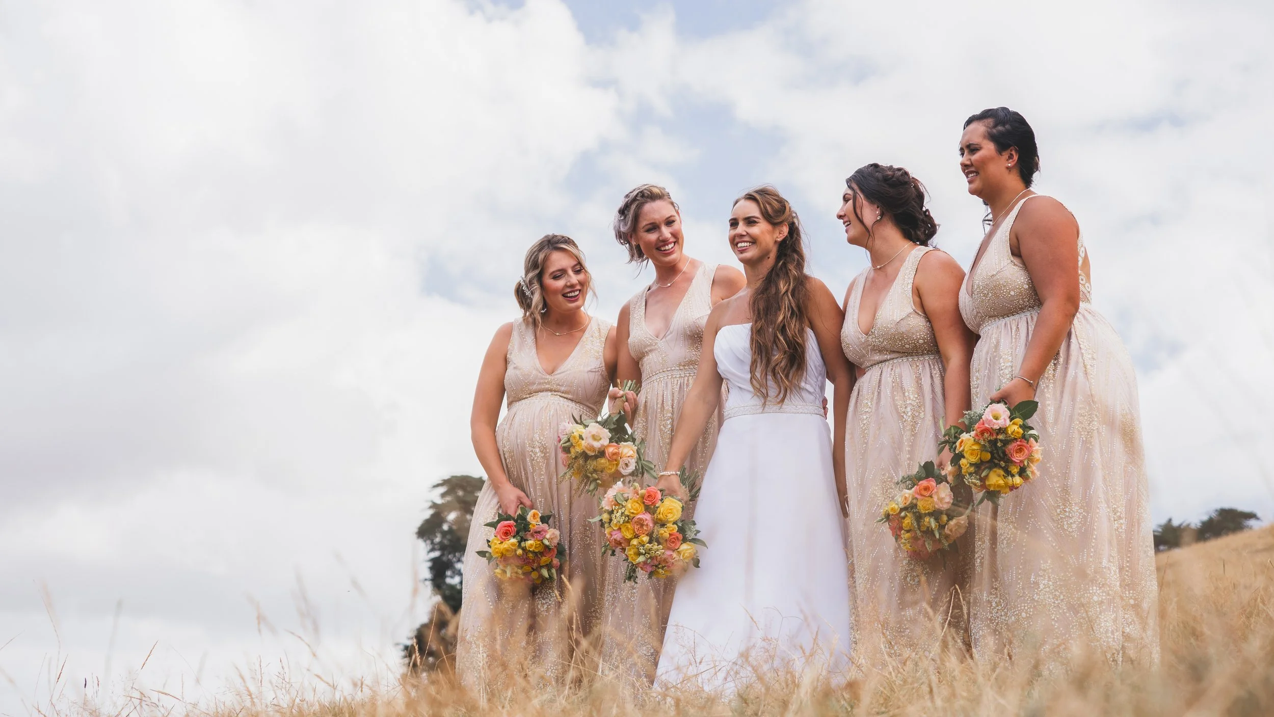 A group of women in wedding dresses holding bouquets, standing outdoors on a cloudy day, smiling and enjoying each other's company.