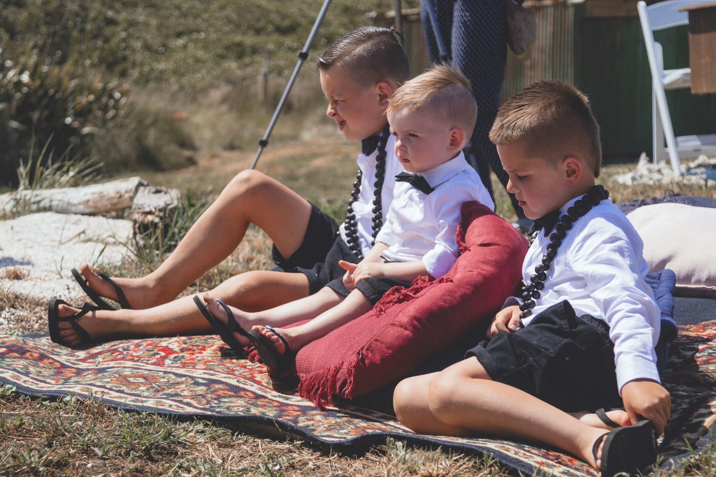 Three young boys seated on a colorful rug outdoors, wearing white shirts and black shorts, with flower necklaces around their necks, participating in a ceremony or ritual on a sunny day.