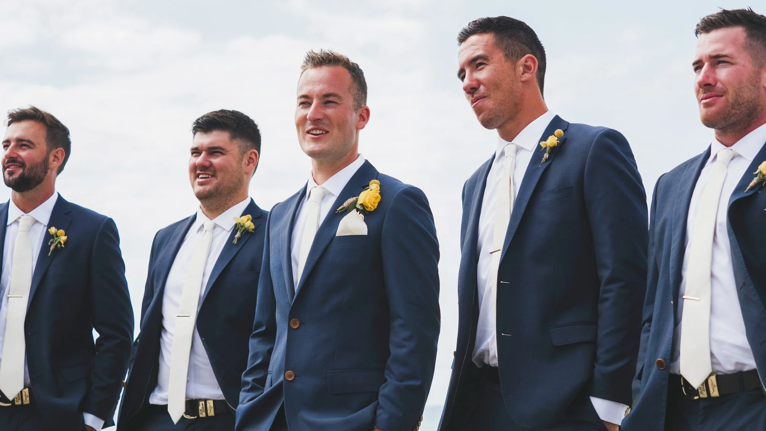 A group of men dressed in navy blue suits with white shirts and white ties, standing outdoors against a cloudy sky.