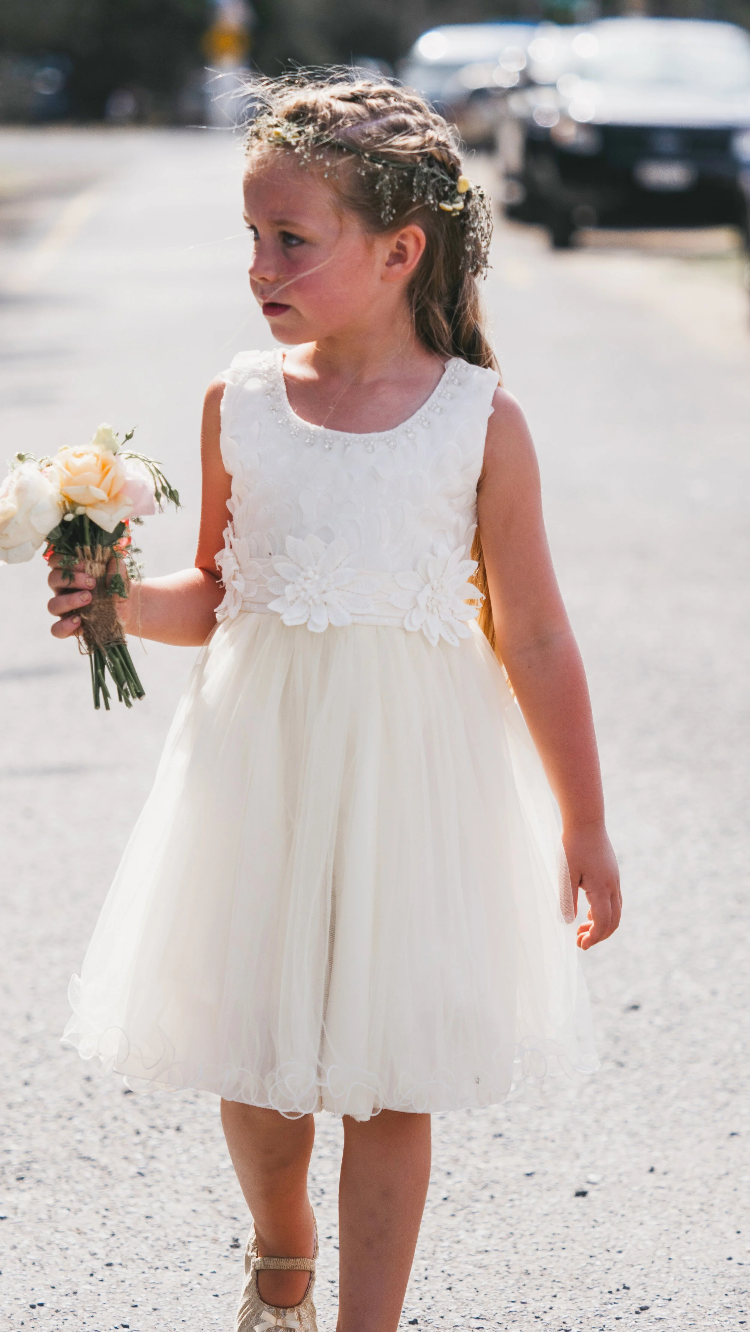 A young girl wearing a white dress with floral embroidery, holding a small bouquet of flowers, standing outdoors on a sunny day.