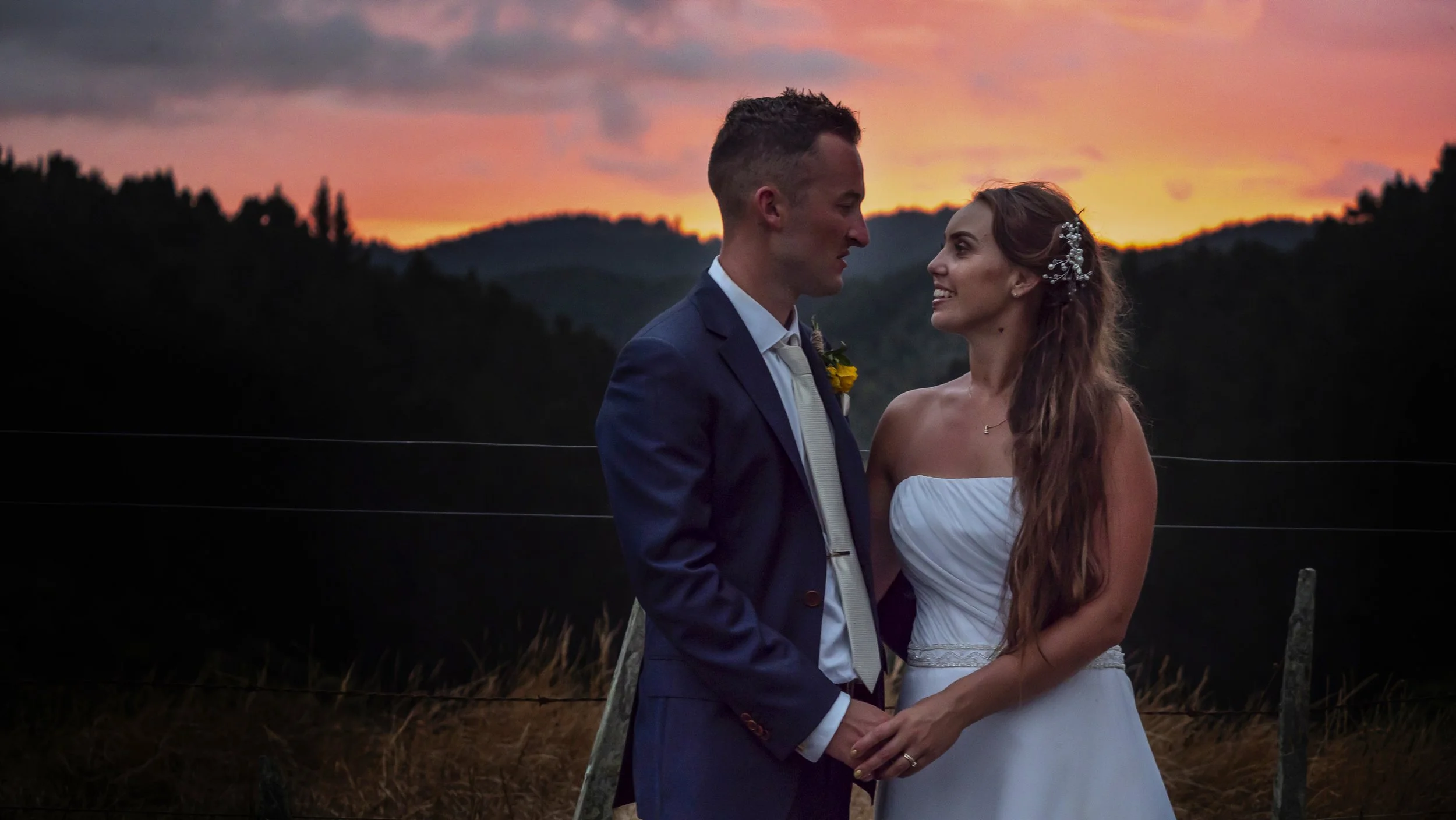 A groom and bride standing close together outdoors during sunset, holding hands and looking into each other's eyes, with a scenic mountain landscape in the background.