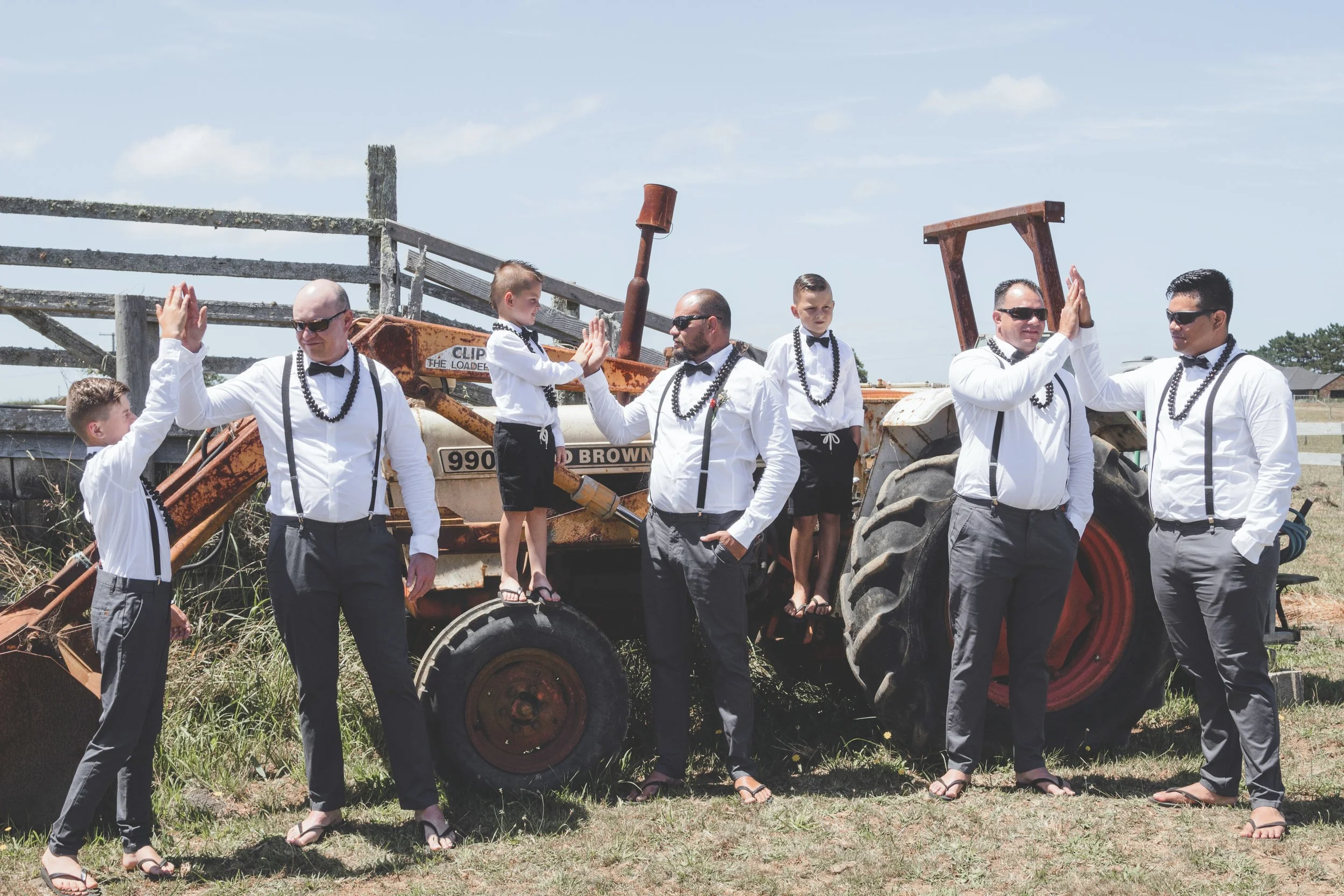 A group of men and boys in formal attire giving high-fives around a rusty orange tractor outdoors on a grassy field.