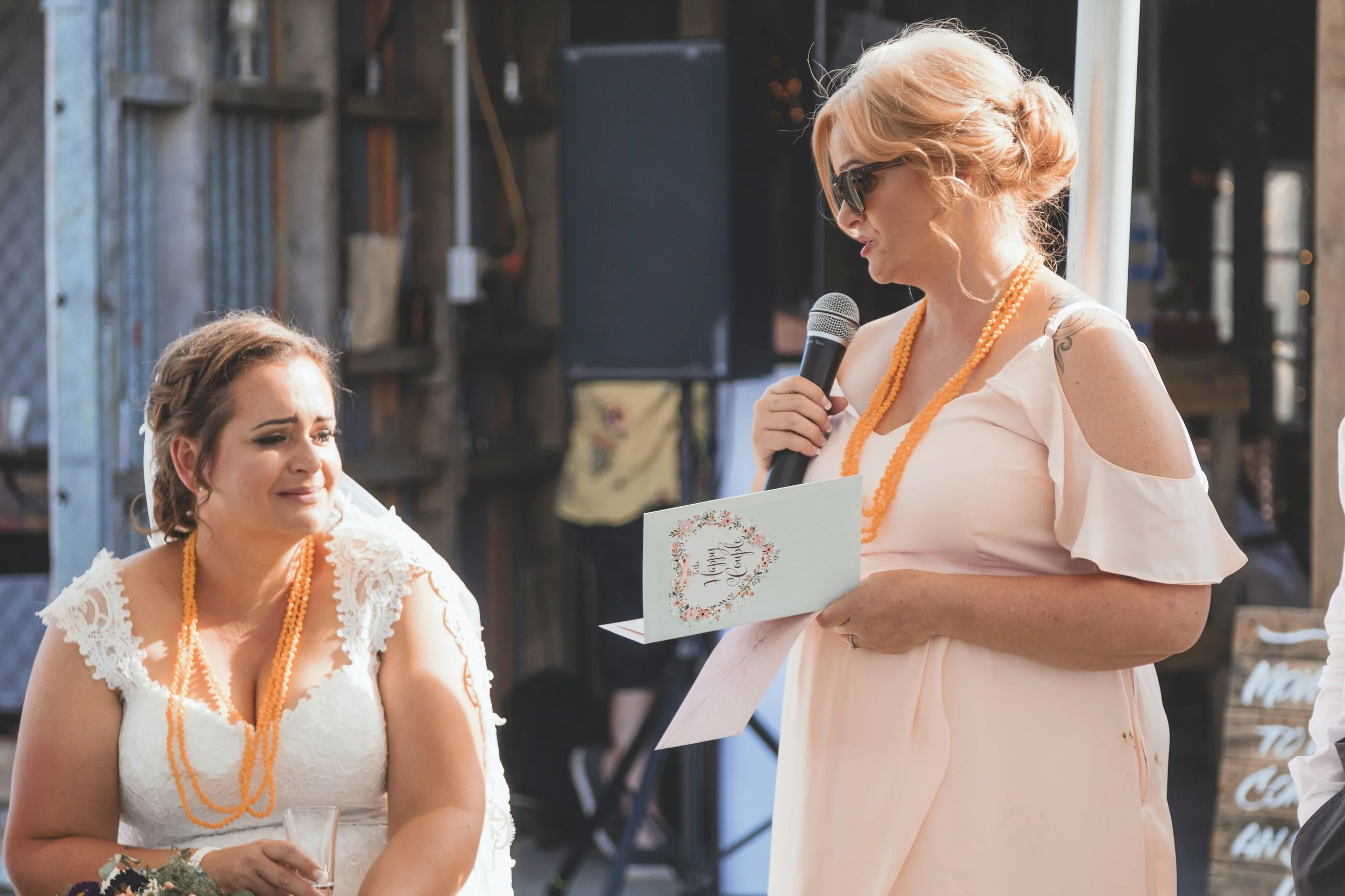 A woman with blonde hair and sunglasses wearing a pink dress and orange beads is giving a speech at an outdoor wedding ceremony, holding a card that says "Happy Wedding". Another woman with brown hair in a white dress with lace and orange beads is sm