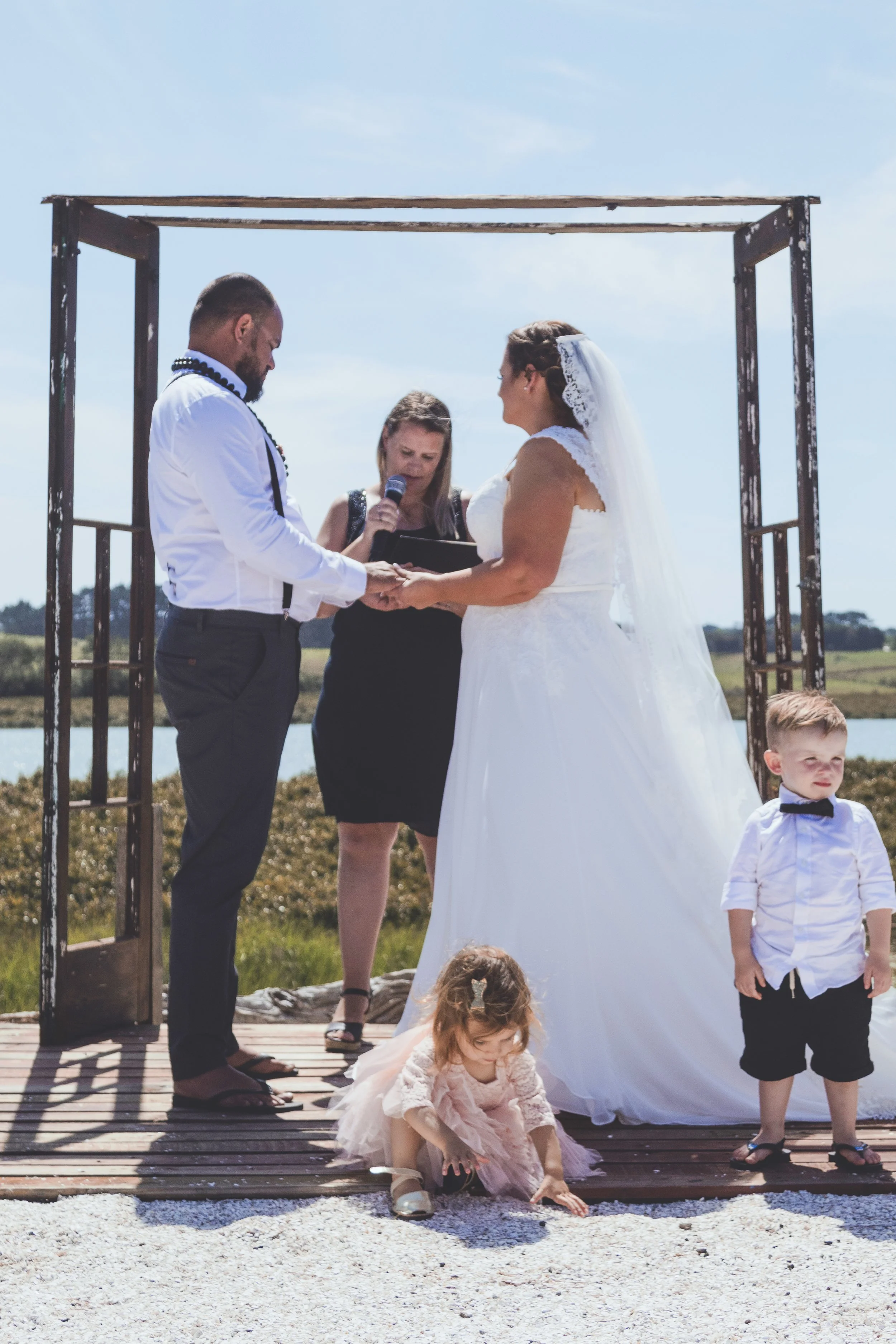 A couple getting married outdoors, kneeling and exchanging vows under a wooden arch, with officiant holding a microphone, and children present, one girl in a pink dress playing on the ground, and a boy in a white shirt and black shorts standing nearb