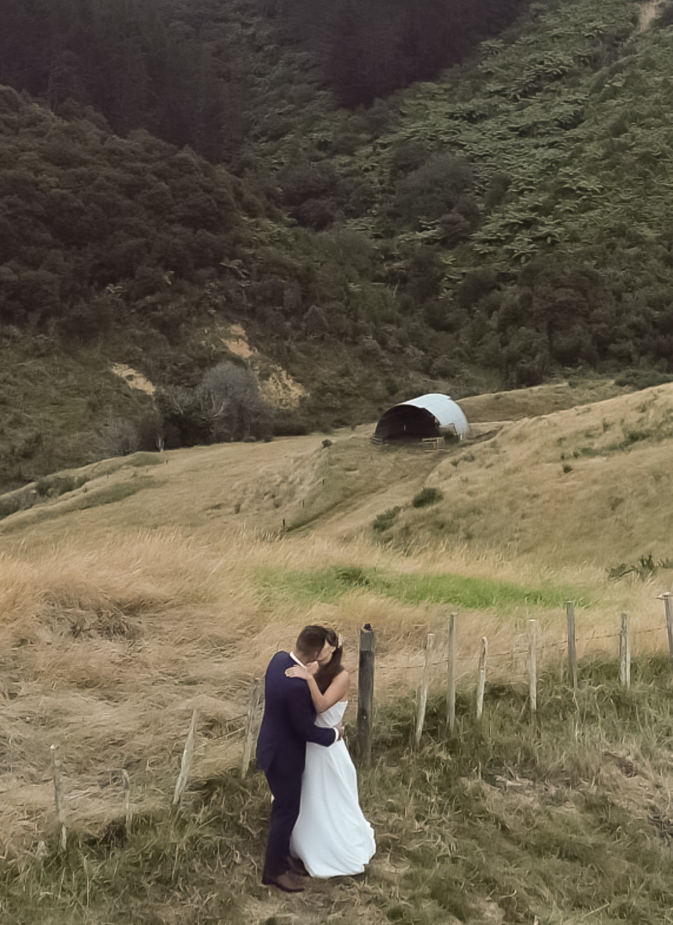 A couple dressed in formal wedding attire sharing an embrace in a grassy, rural landscape near a fence with mountains and a barn or storage building in the background.
