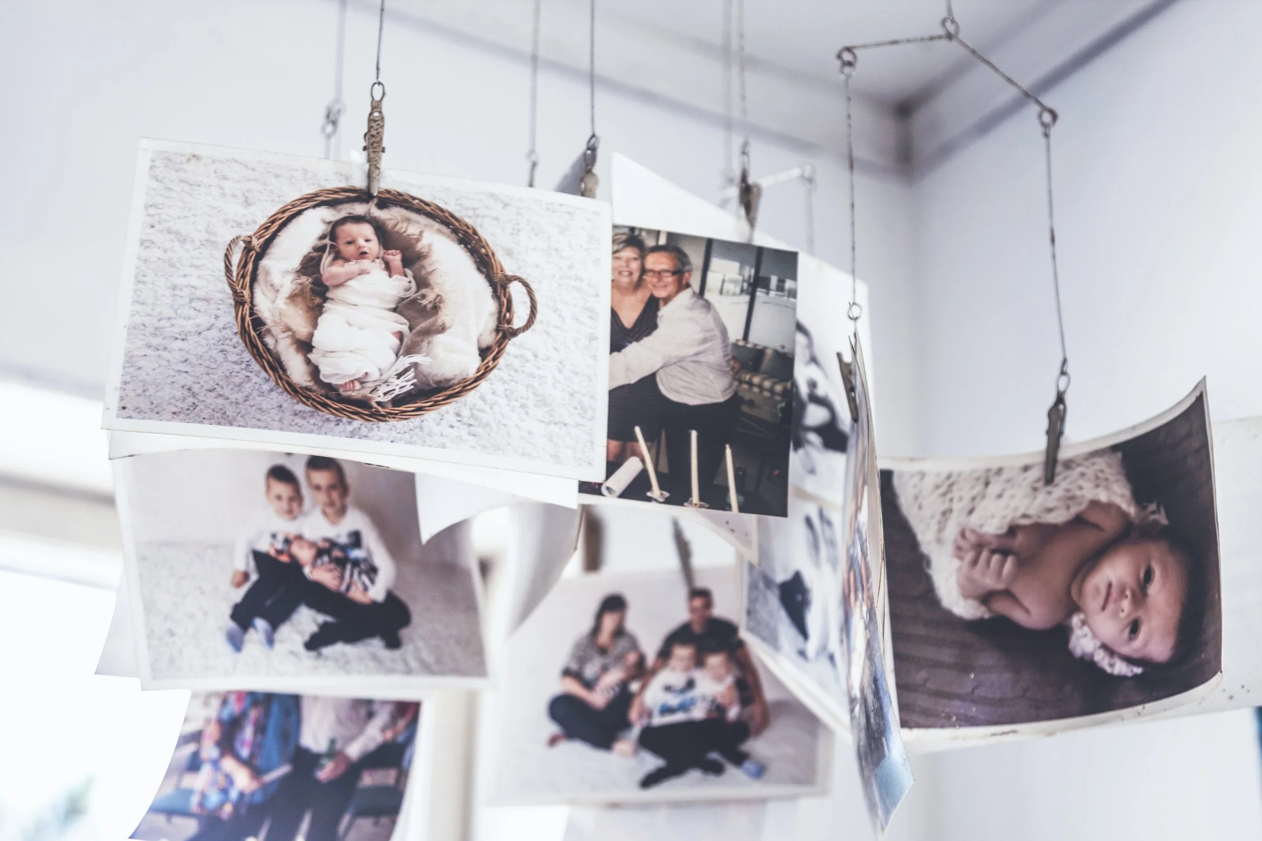 Photographs hanging on wires displaying various family moments, including a baby in a basket, children playing, and a couple sitting together, with some partially out of frame.
