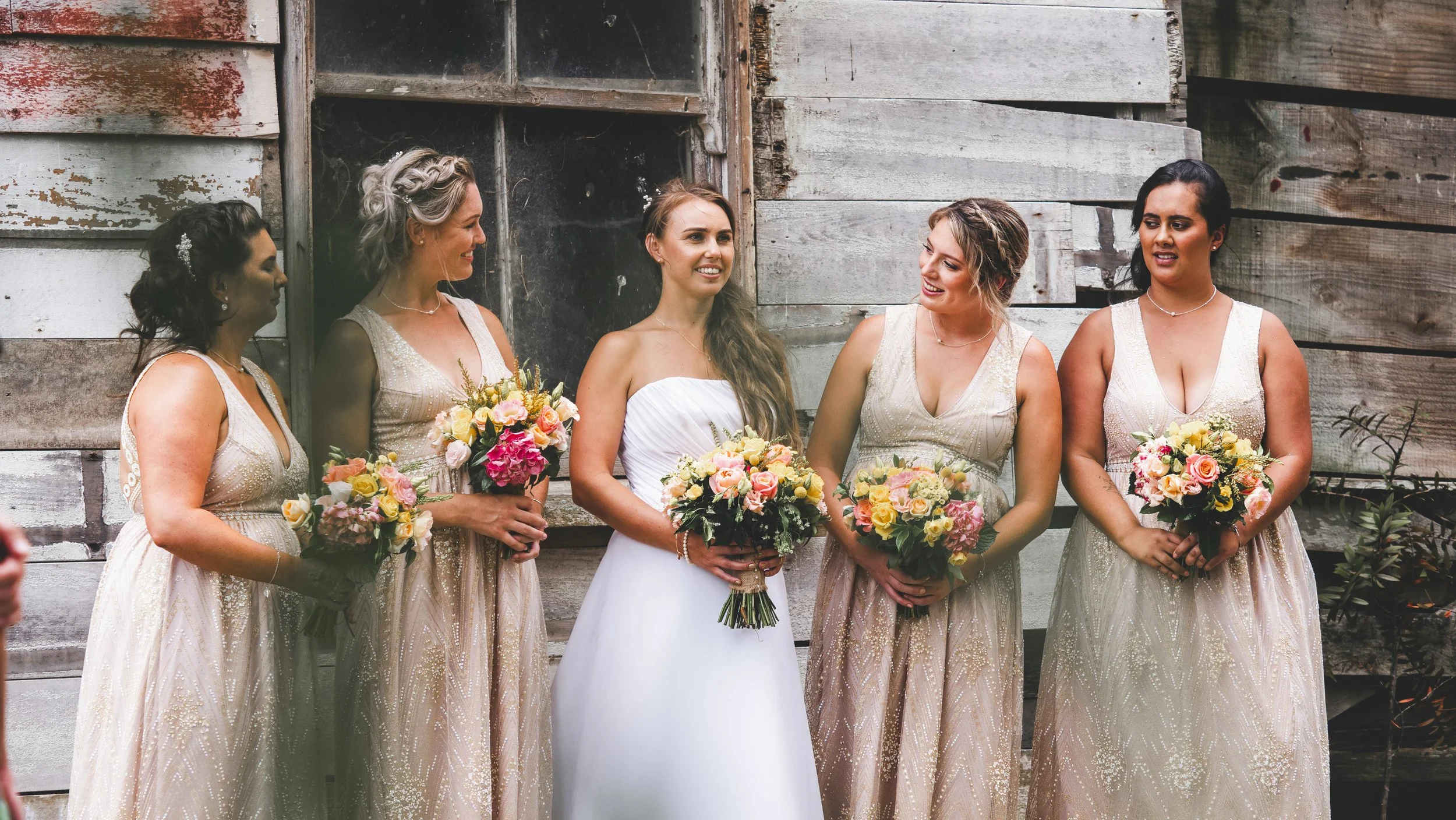 A bride and five bridesmaids standing outside in front of a weathered wooden wall, all holding bouquets of flowers, during a wedding celebration.