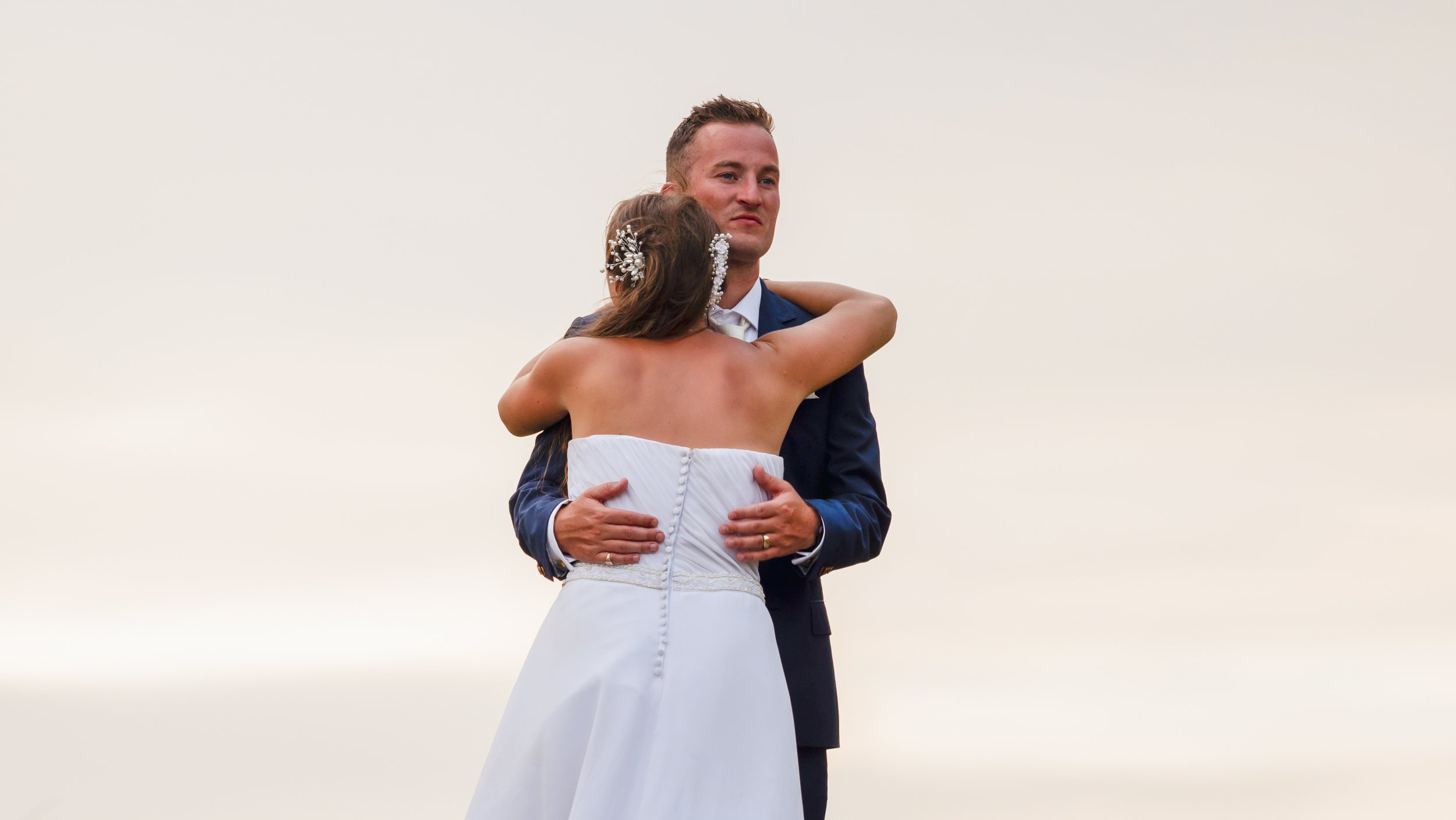 A couple, dressed in wedding attire, embracing outdoors against a plain sky background. The woman wears a strapless white wedding dress with decorative hair accessories, and the man wears a dark suit with a white shirt.
