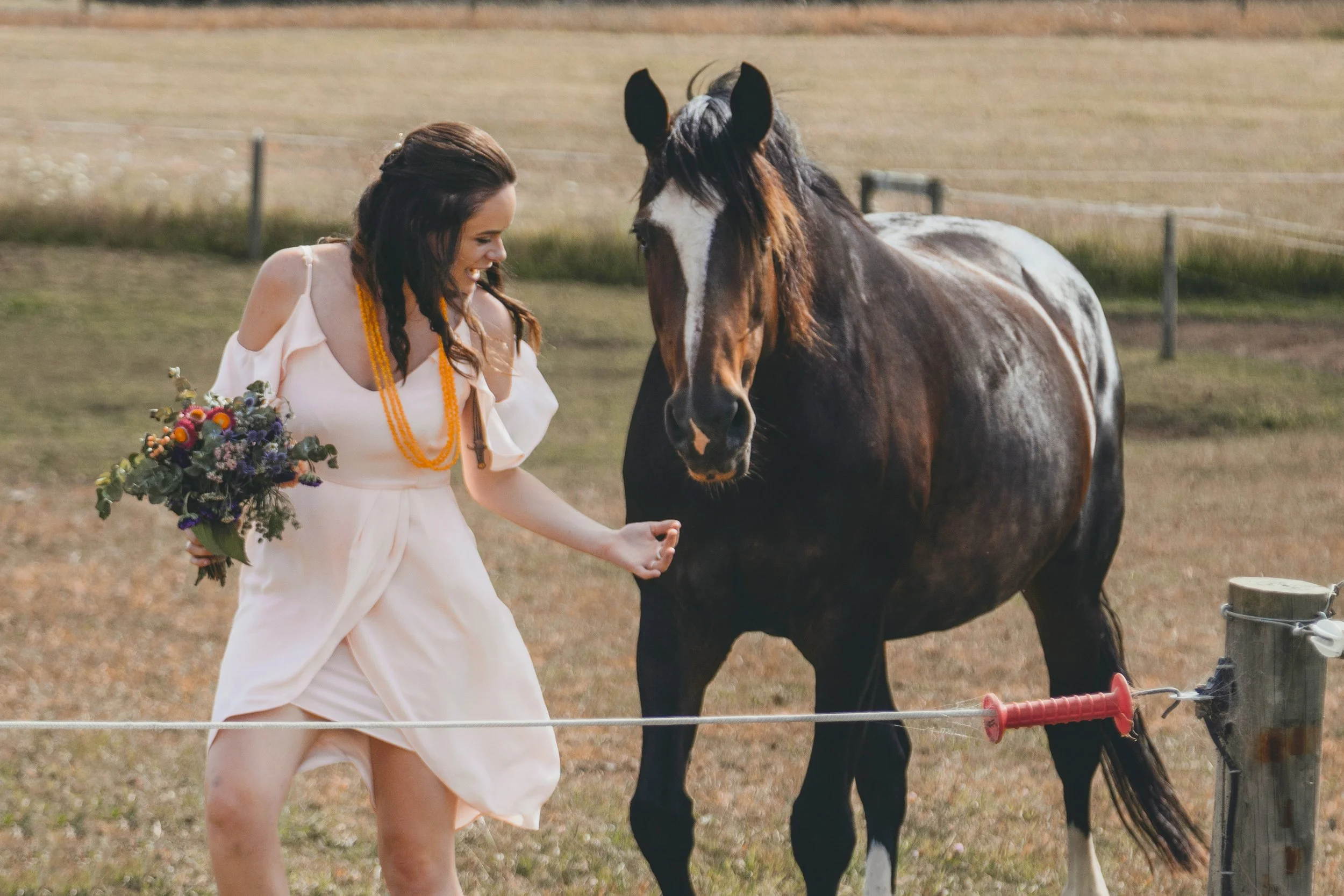 A woman in a white dress holding a bouquet of flowers standing next to a brown and white horse in a field.