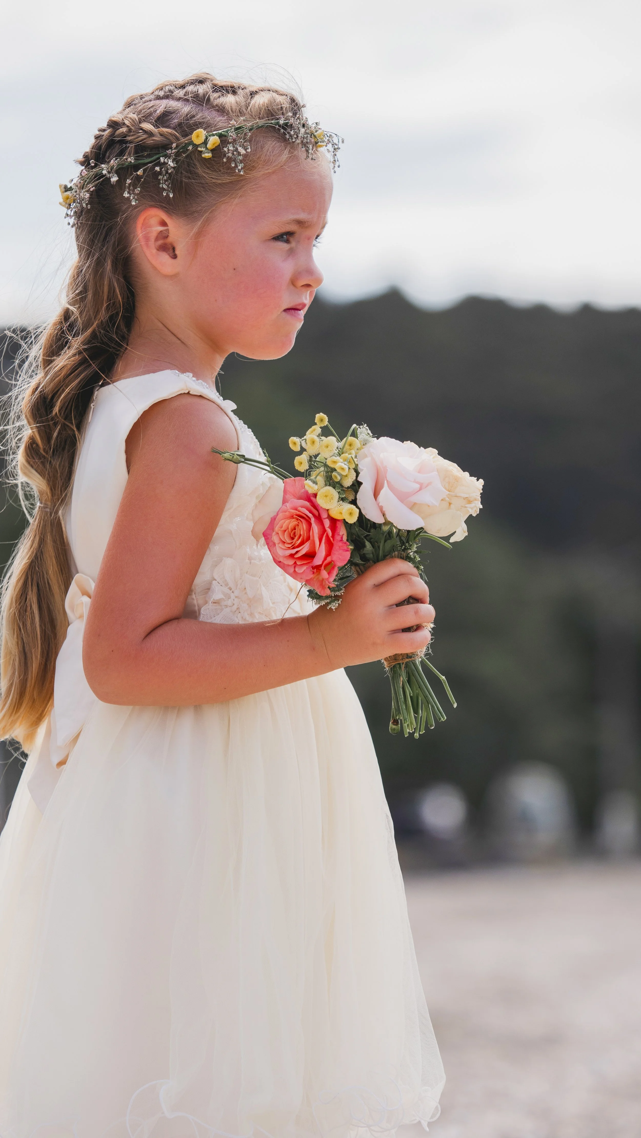 A young girl wearing a cream-colored dress, with a floral crown in her braided hair, holding a bouquet of pink, white, and yellow flowers, standing outdoors with a blurred landscape background.