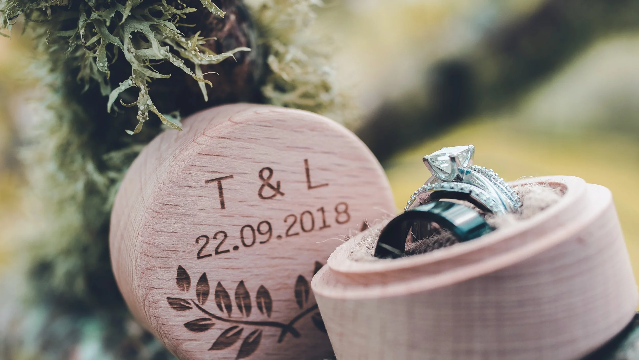 A pink wooden box with engraved initials 'T & L', a date '22.09.2018', and a decorative leaf design, surrounded by green moss, a bouquet of pink roses, and wedding rings, including an engagement ring with a large diamond.