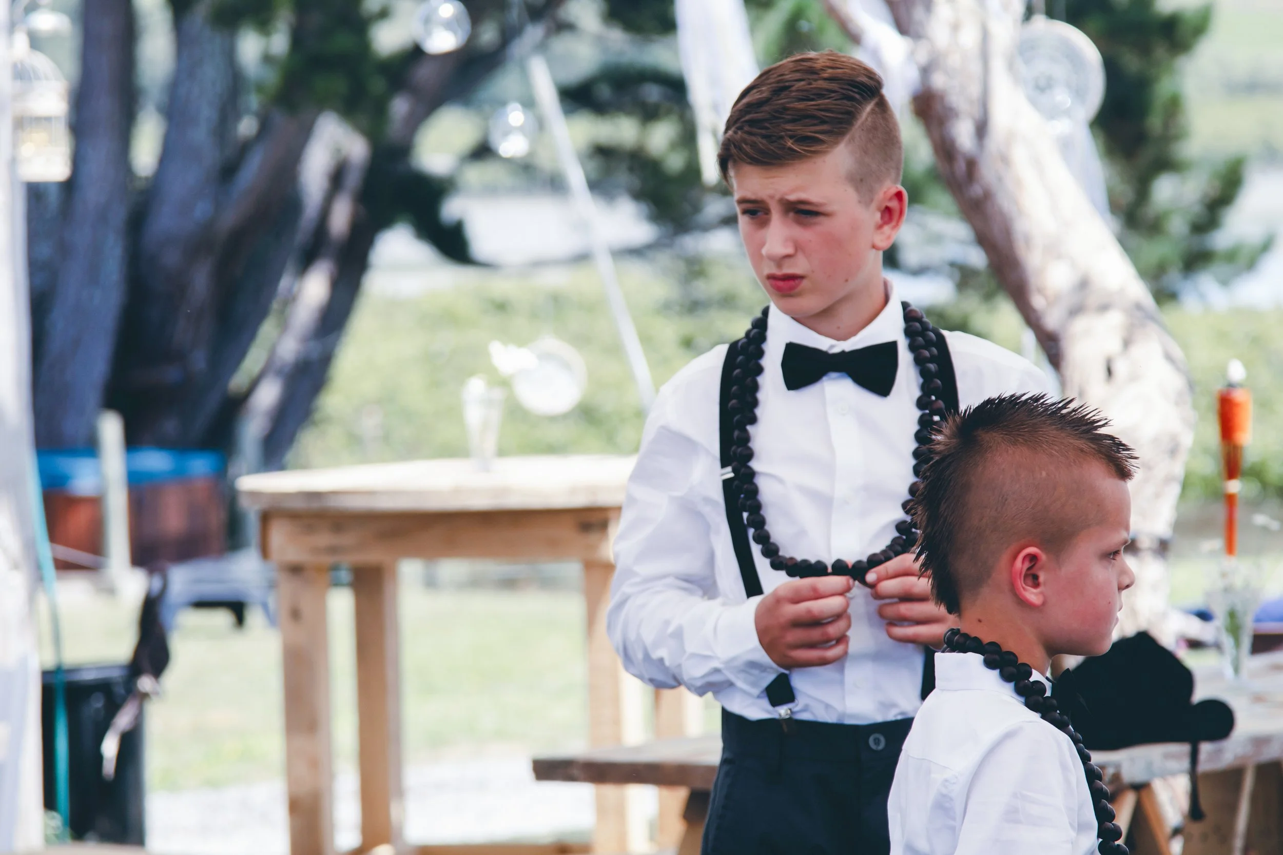A young boy in a tuxedo with a bow tie and beaded necklace stands outdoors, looking to his left with a serious expression. Other children are visible around him, and the background includes trees and hanging lanterns.