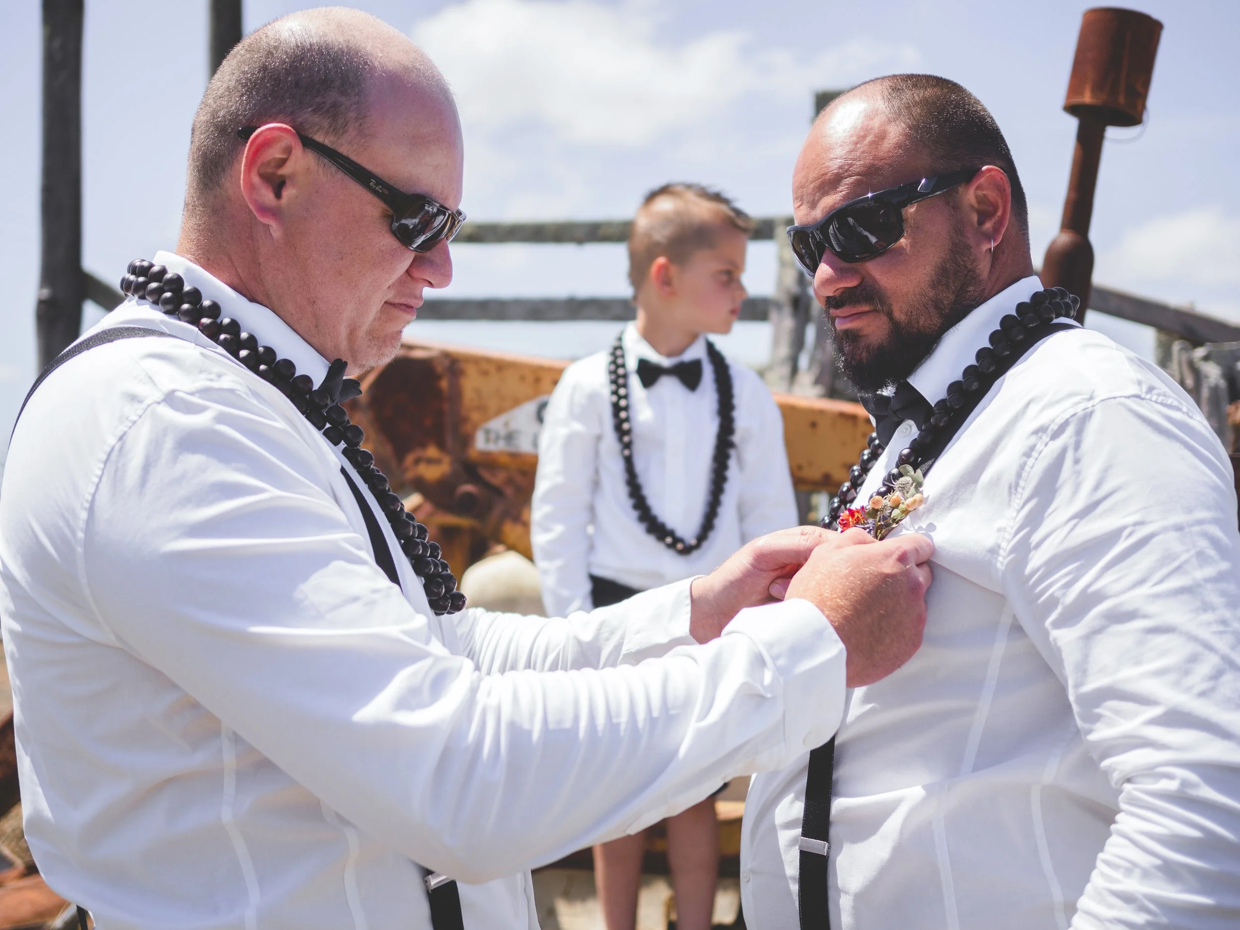 Two men in white shirts and black beaded necklaces, wearing sunglasses, are exchanging rings outdoors on a sunny day, with a young boy standing behind them.