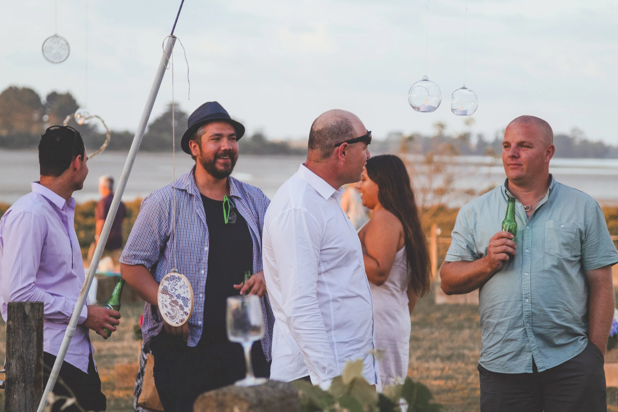 Group of people socializing outdoors near the beach during sunset, with candles in glass holders hanging above.