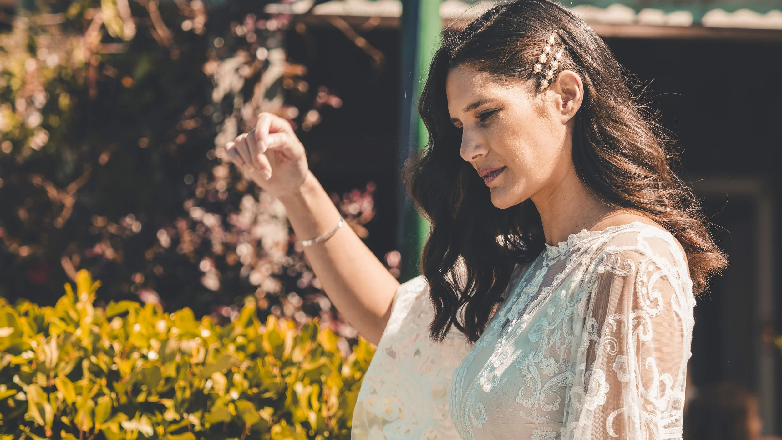 A woman with long dark hair, wearing a white lace dress, is touching a yellow flower bush in a garden on a sunny day.