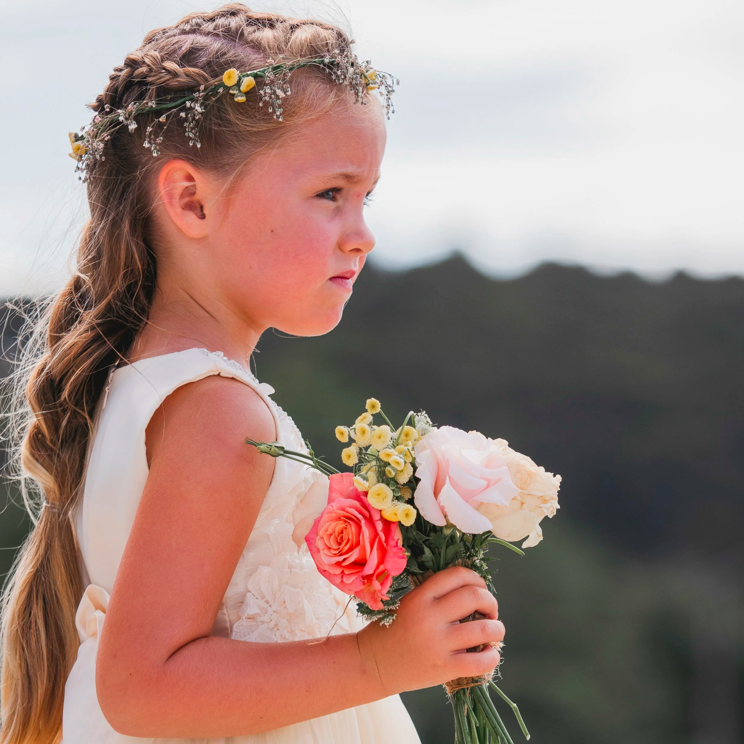 A young girl with long hair in braids, wearing a floral crown, holding a bouquet of pink, white, and yellow flowers against a blurred outdoor background.