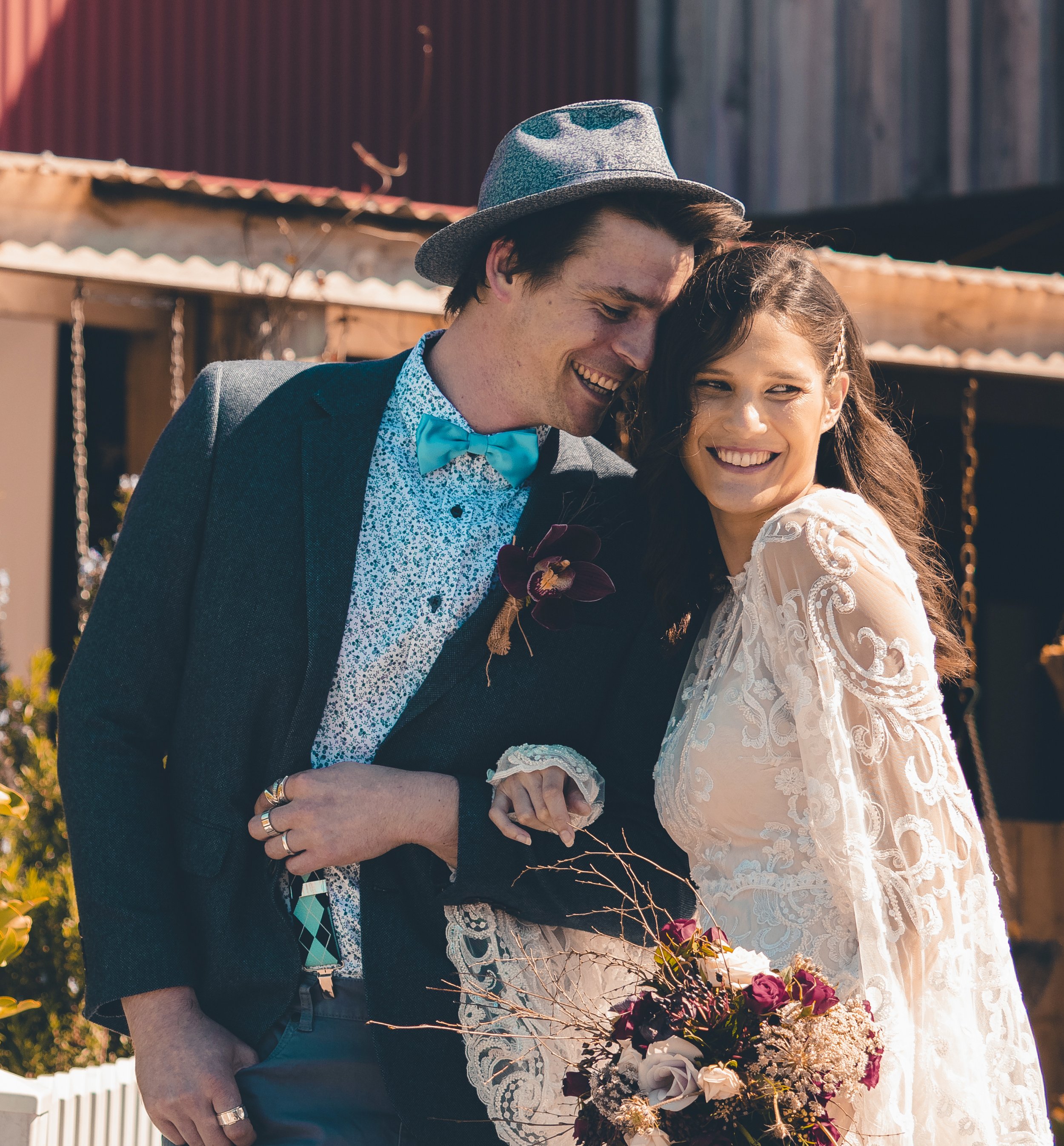 A young couple at a wedding, smiling with their heads close together, holding a bouquet of flowers, outdoors with a wooden building in the background.