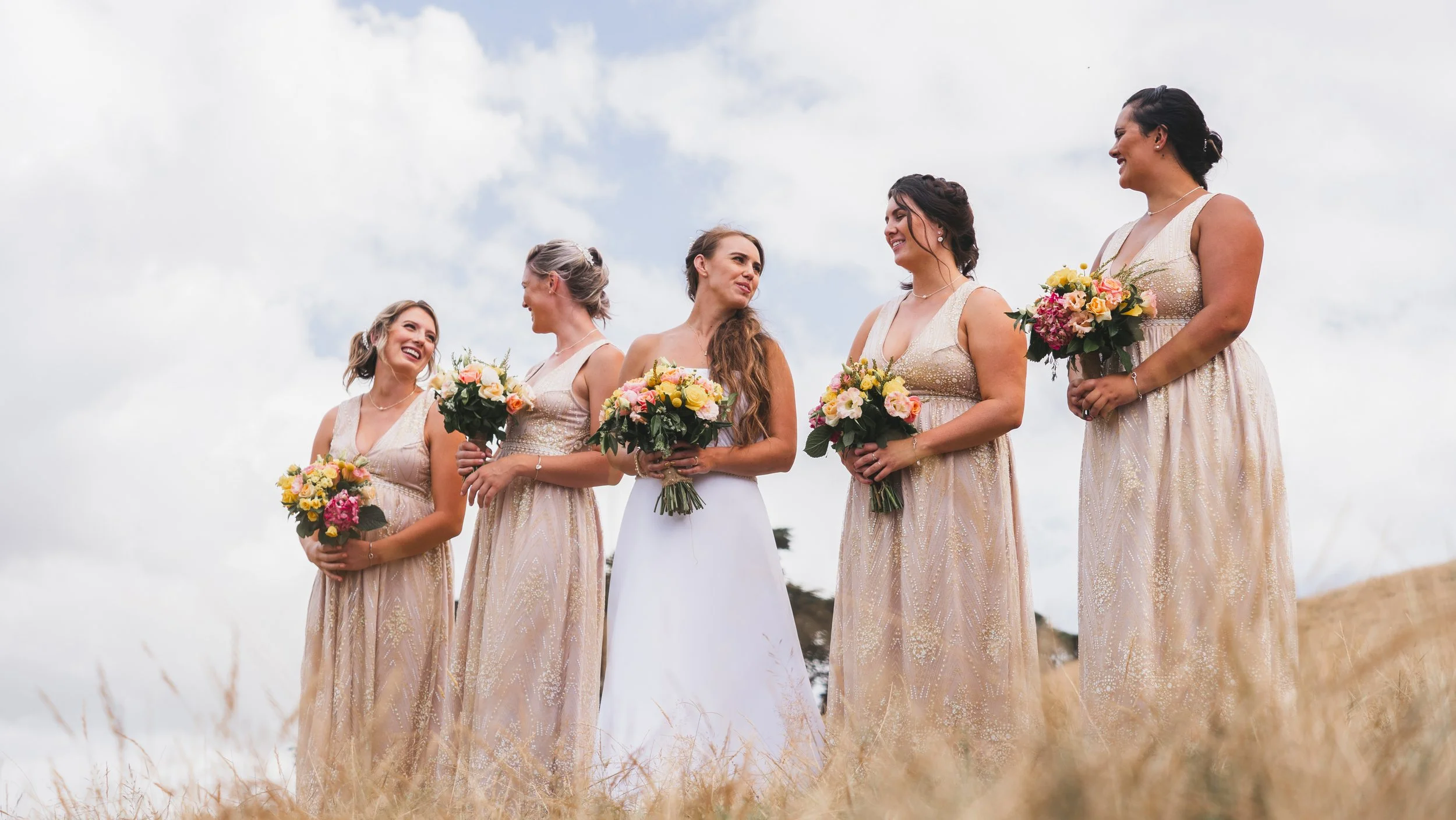 A bride and her four bridesmaids standing outdoors, holding bouquets of flowers, smiling and talking, with a cloudy sky in the background.