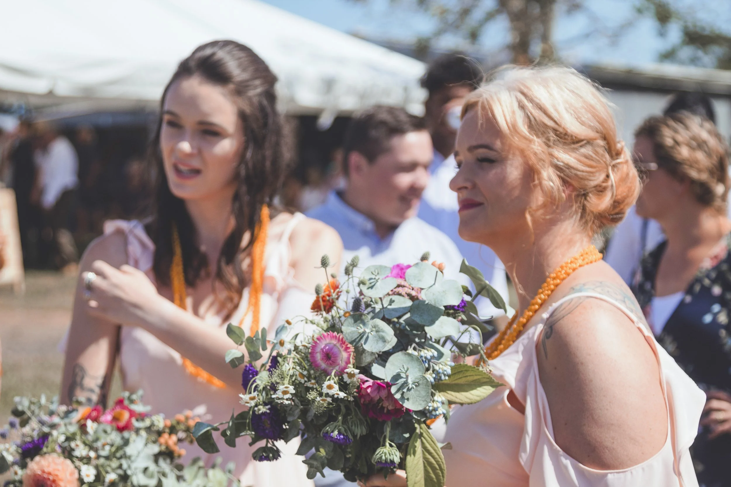 Women at an outdoor event exchanging flowers, wearing casual summer clothes with balloons and tents in the background.