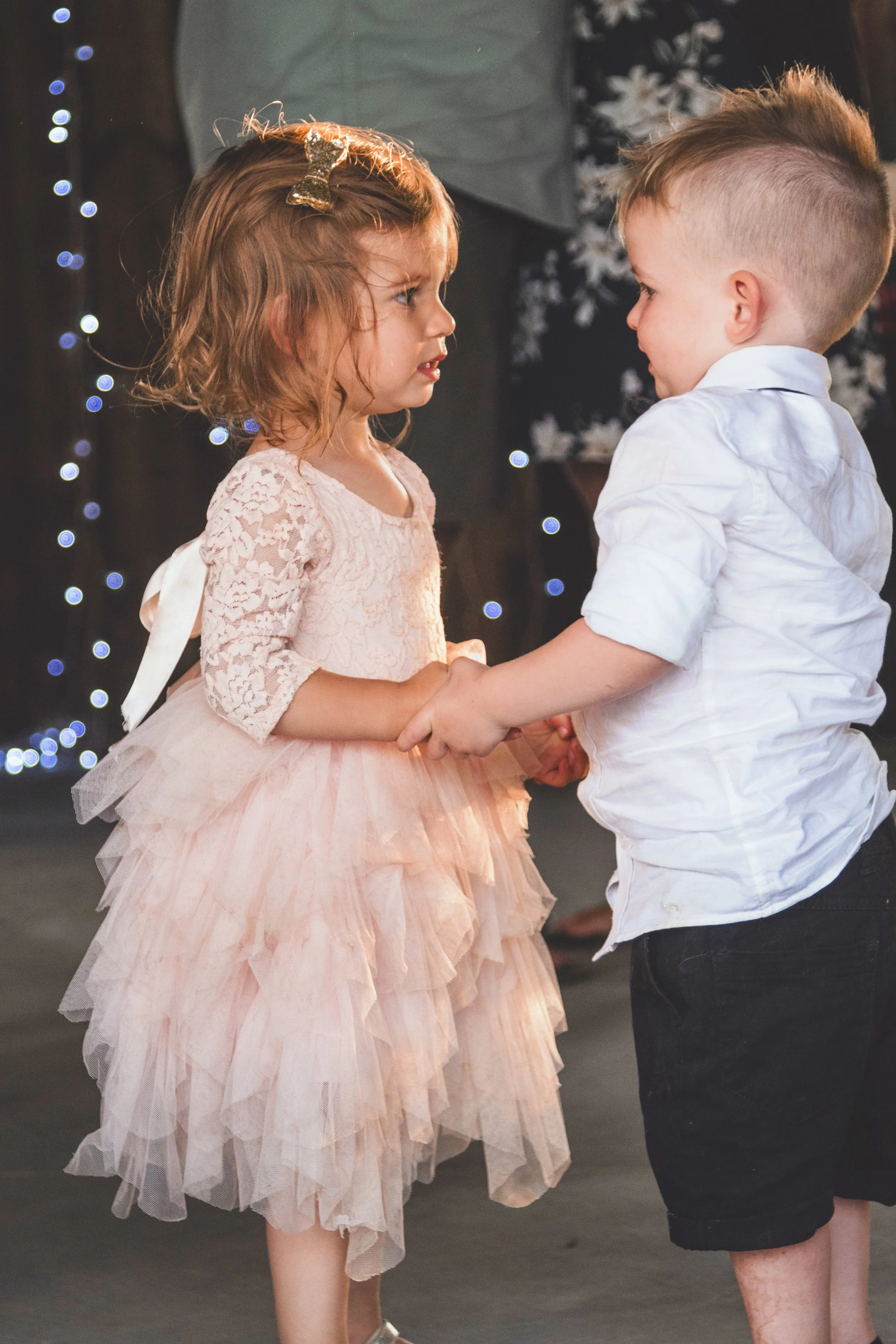 A young girl and boy holding hands and facing each other indoors with background Christmas lights and decorations.
