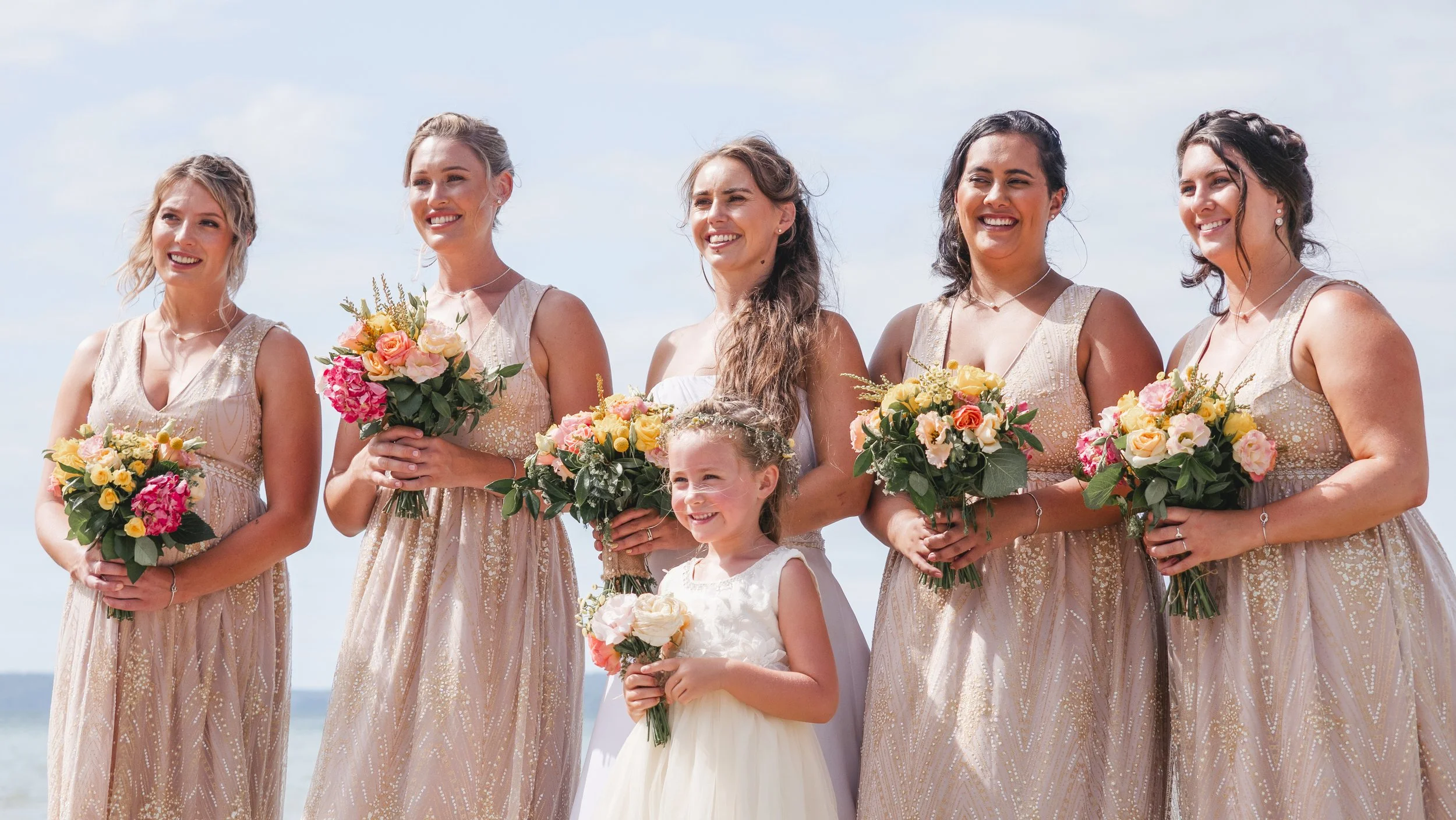 A group of women and a girl at a wedding, holding colorful bouquets of flowers, standing outdoors with a cloudy sky in the background.