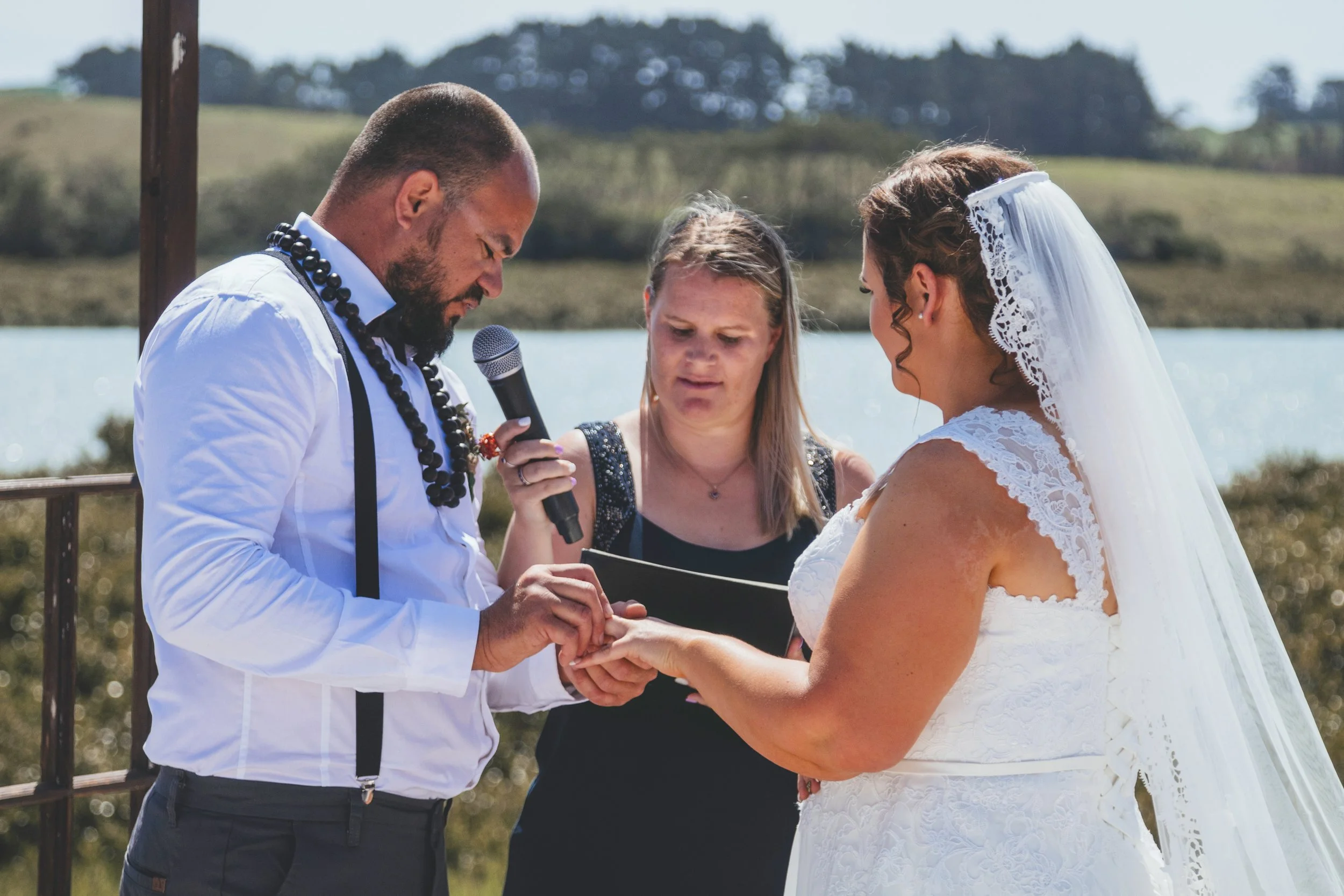 A couple getting married outdoors by a lake, with a woman officiating. The groom is holding the bride's hand as they exchange vows.