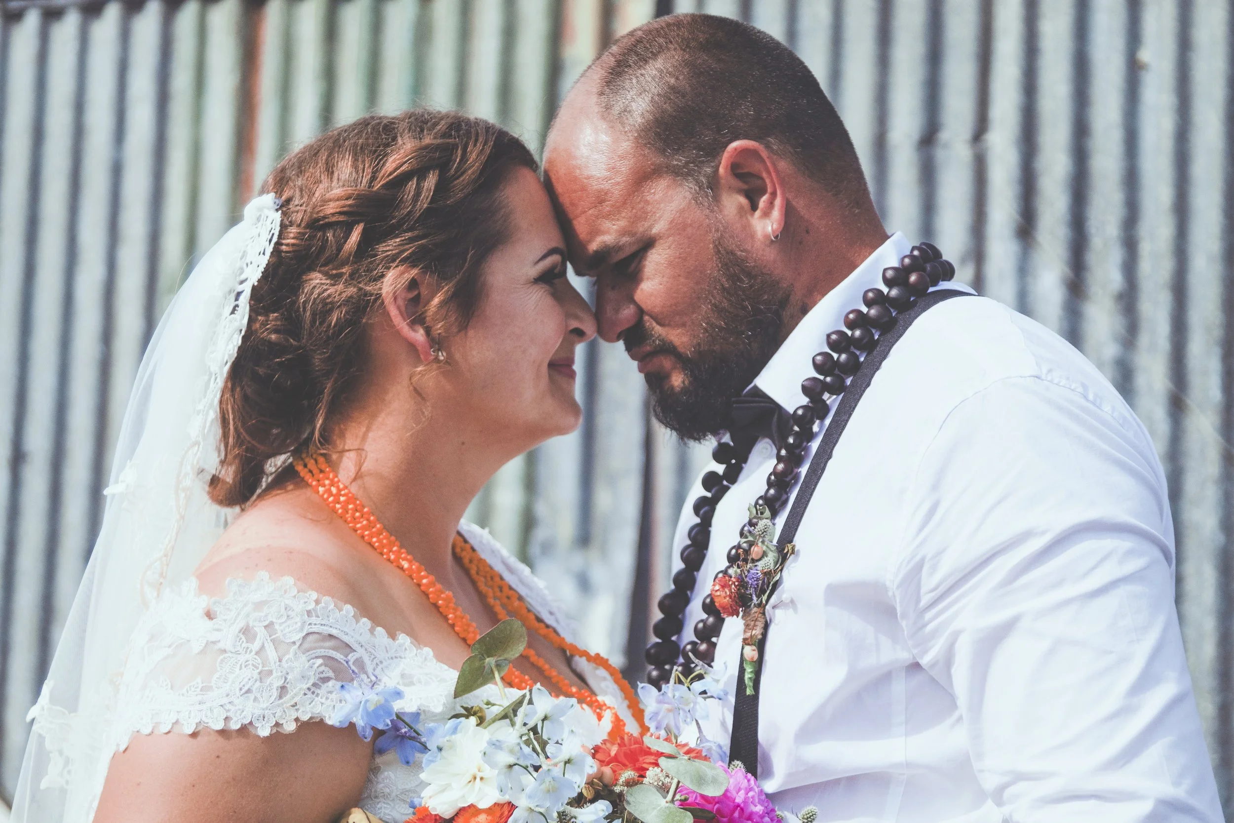 A bride and groom touching foreheads during their wedding ceremony, both smiling softly and holding a bouquet of flowers. The bride has brown hair styled in an updo with a lace veil, and the groom has a shaved head, beard, and wears a white shirt wit