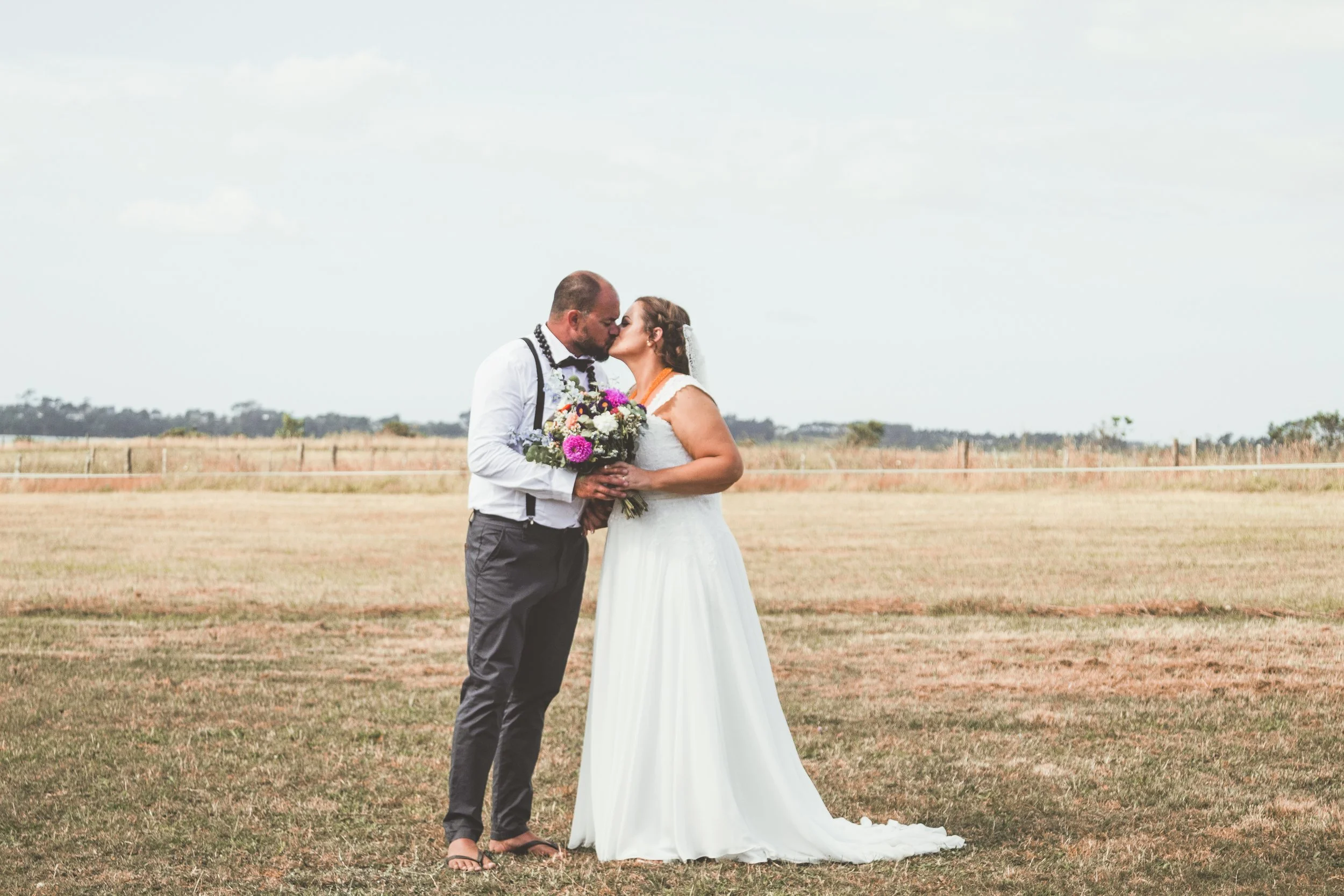 A bride and groom share a kiss outdoors on a grassy field during their wedding, with the groom holding a bouquet of flowers and wearing a white shirt with suspenders, and the bride in a white wedding gown.