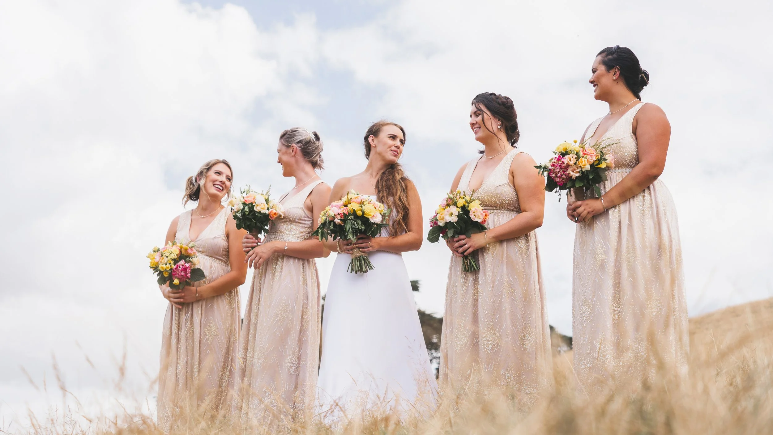 A bride and five bridesmaids standing outdoors, each holding a bouquet of flowers, wearing light pink dresses, with a cloudy sky in the background.